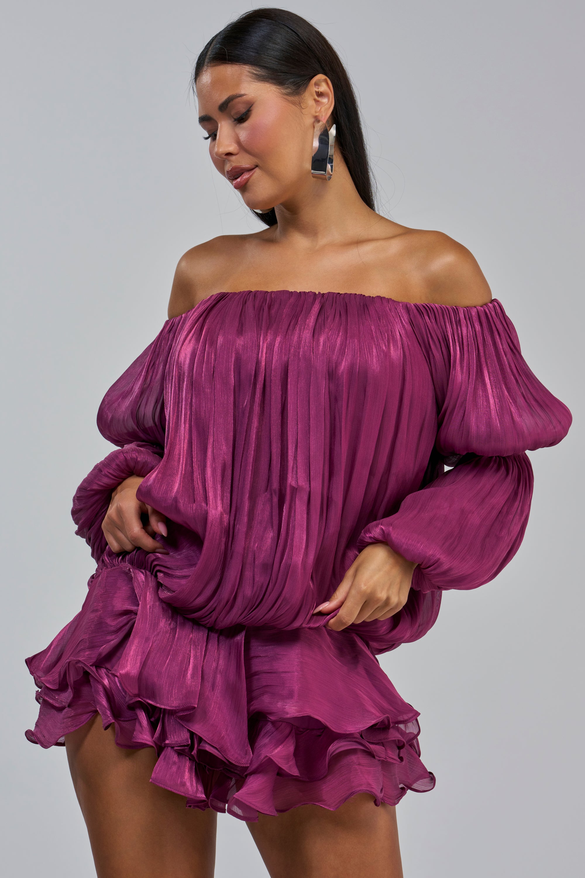 A woman with long dark hair and statement earrings is smiling and looking down while wearing the FAVORITE DAUGHTER CHIFFON MINI DRESS IN PURPLE, featuring a shiny off-the-shoulder design with voluminous sleeves and a ruffled skirt.