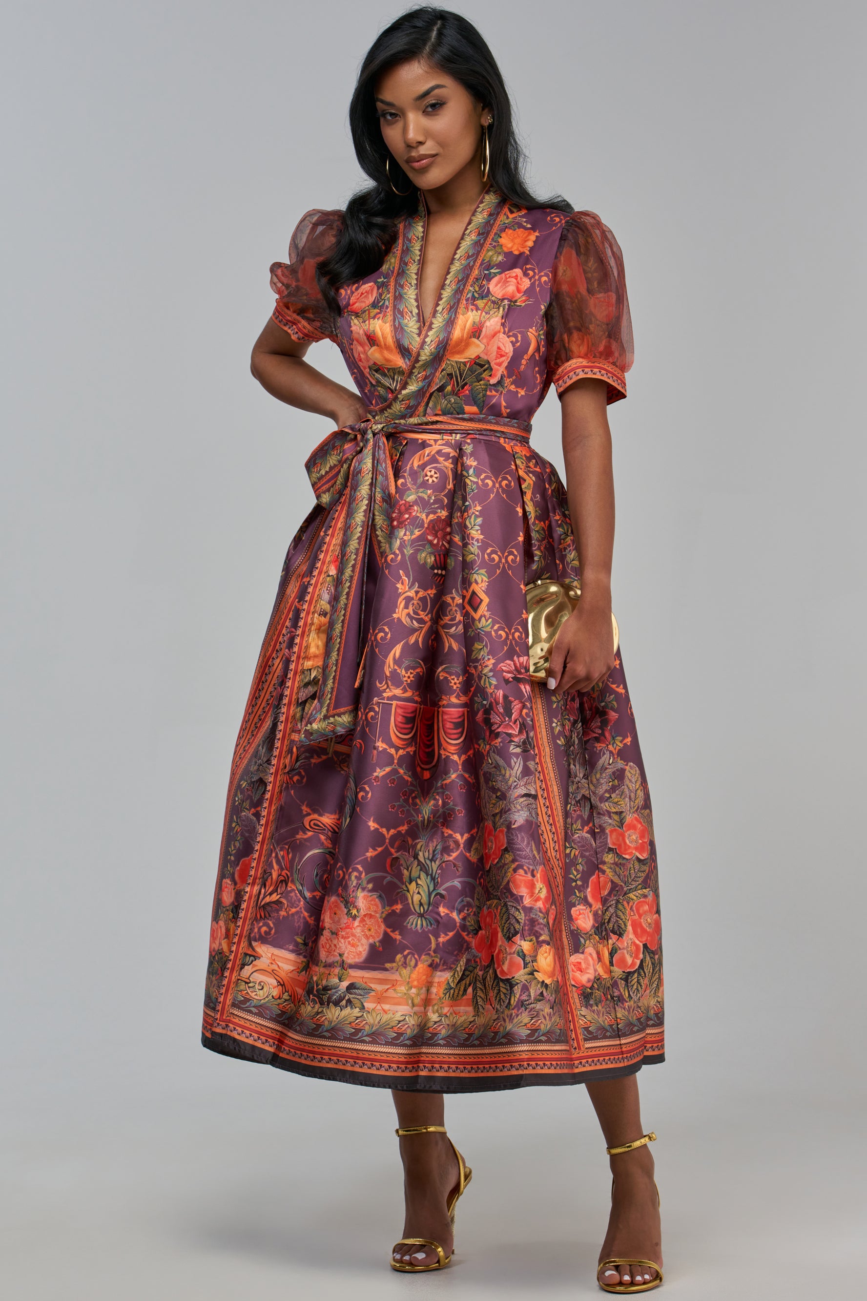 A person wears a FLOWER GIRL SATIN SHORT SLEEVE MIDI DRESS IN RUST COMBO with short sheer sleeves, a v-neckline, and an ornate pattern, paired with gold strappy heels and a gold clutch.