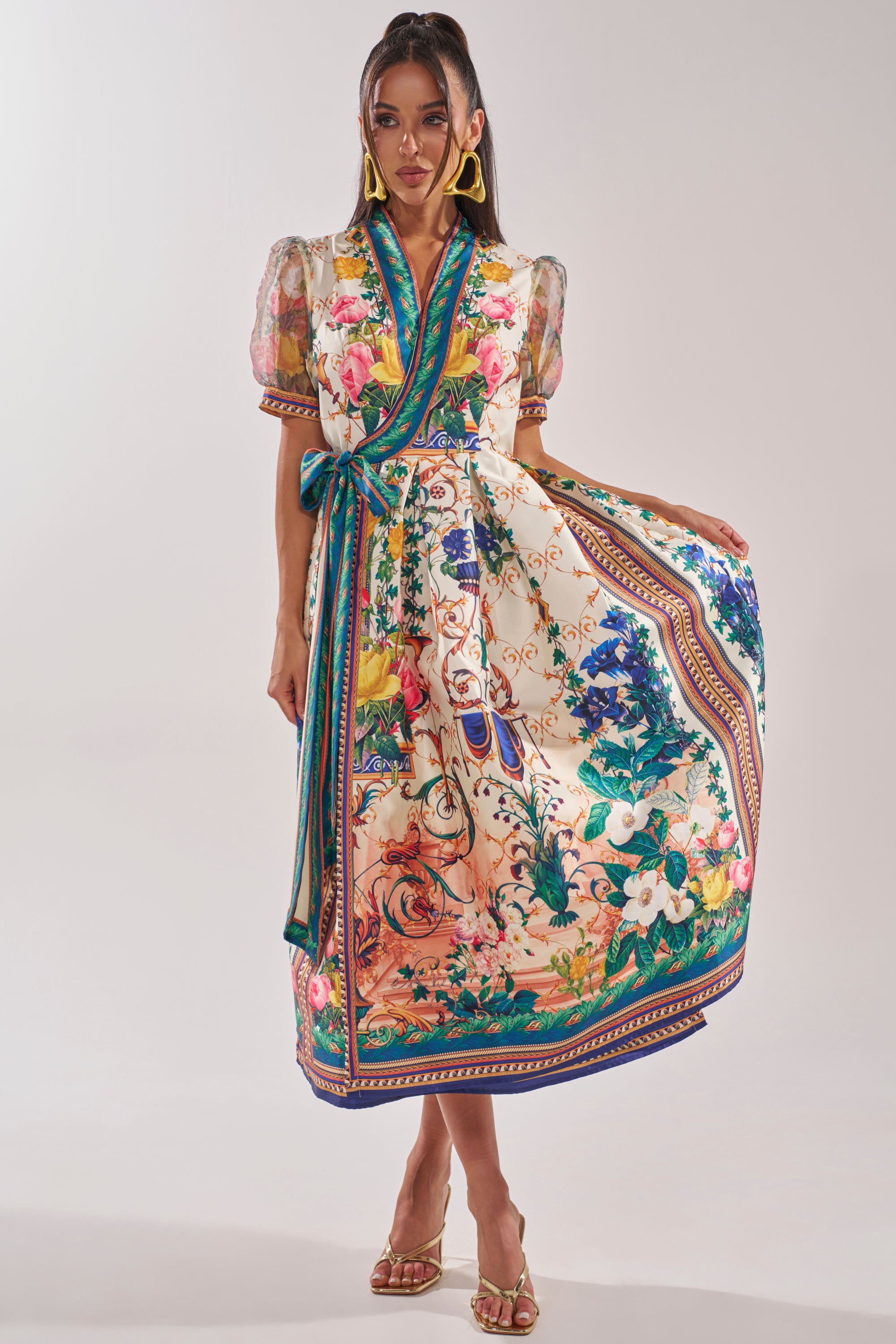 A woman wears the FLOWER GIRL SATIN MAXI DRESS, a vibrant floral wrap maxi with puff sleeves and a blue sash. She pairs it with gold heels, statement earrings, and gracefully holds out part of her skirt.