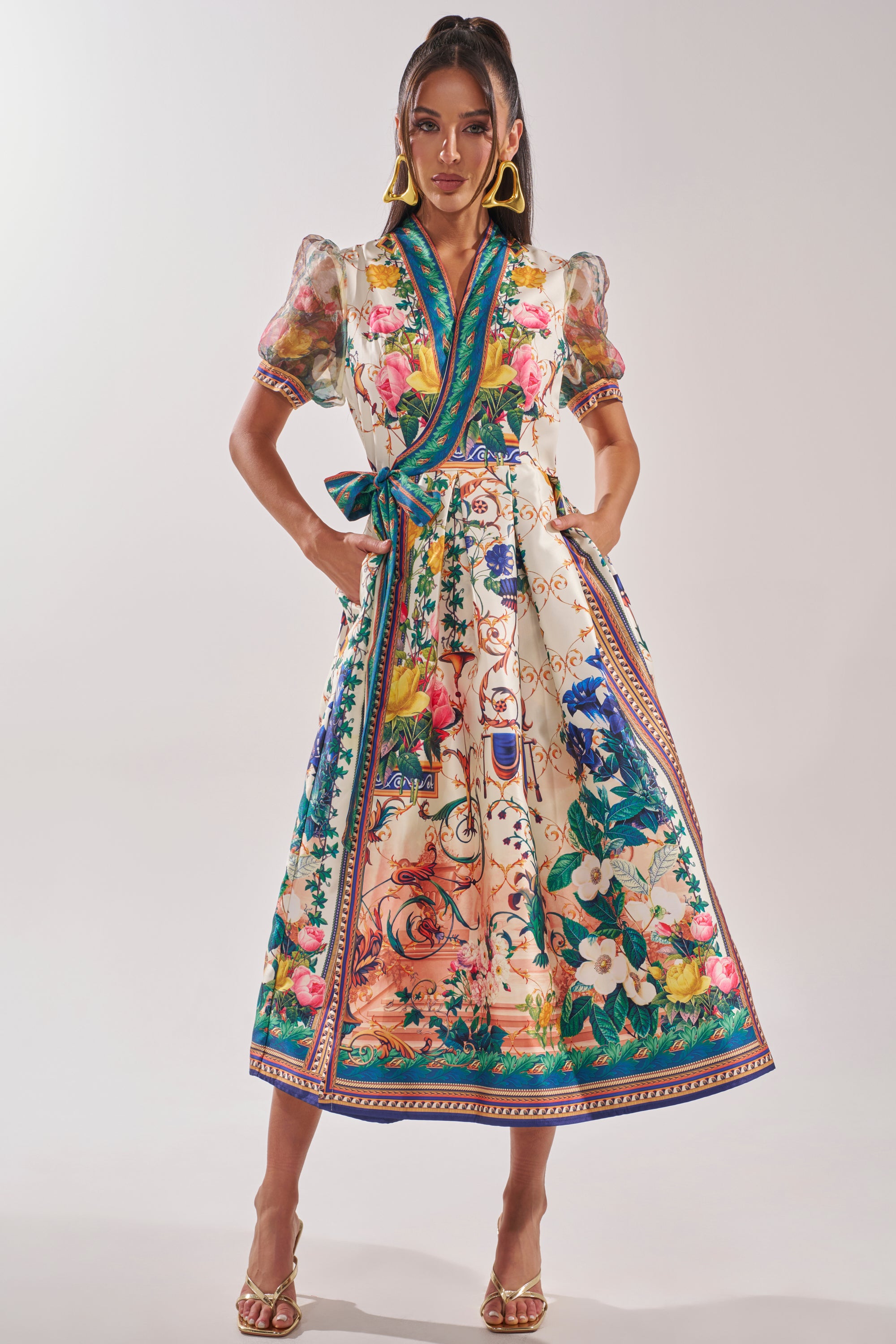 A woman stands confidently in the FLOWER GIRL SATIN MAXI DRESS, a botanical print wrap maxi with puff sleeves and a waist sash. She completes the look with gold statement earrings, matching gold heels, and wears her long dark hair down. 1