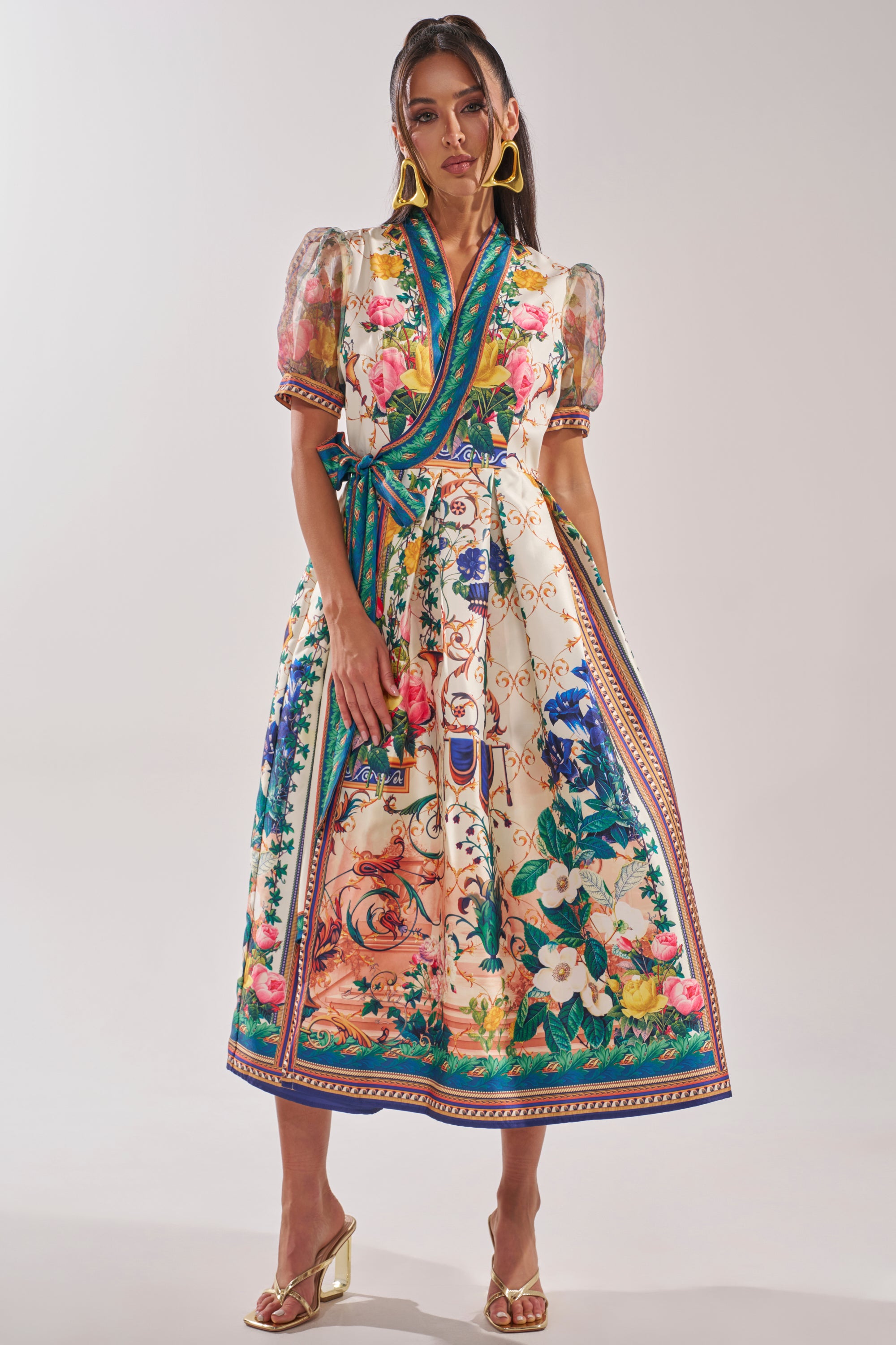 A woman in the FLOWER GIRL SATIN MAXI DRESS, featuring a colorful botanical print, puff sleeves, and sash tie, stands against a plain background. She wears gold earrings, gold heeled sandals, and has long hair. 2