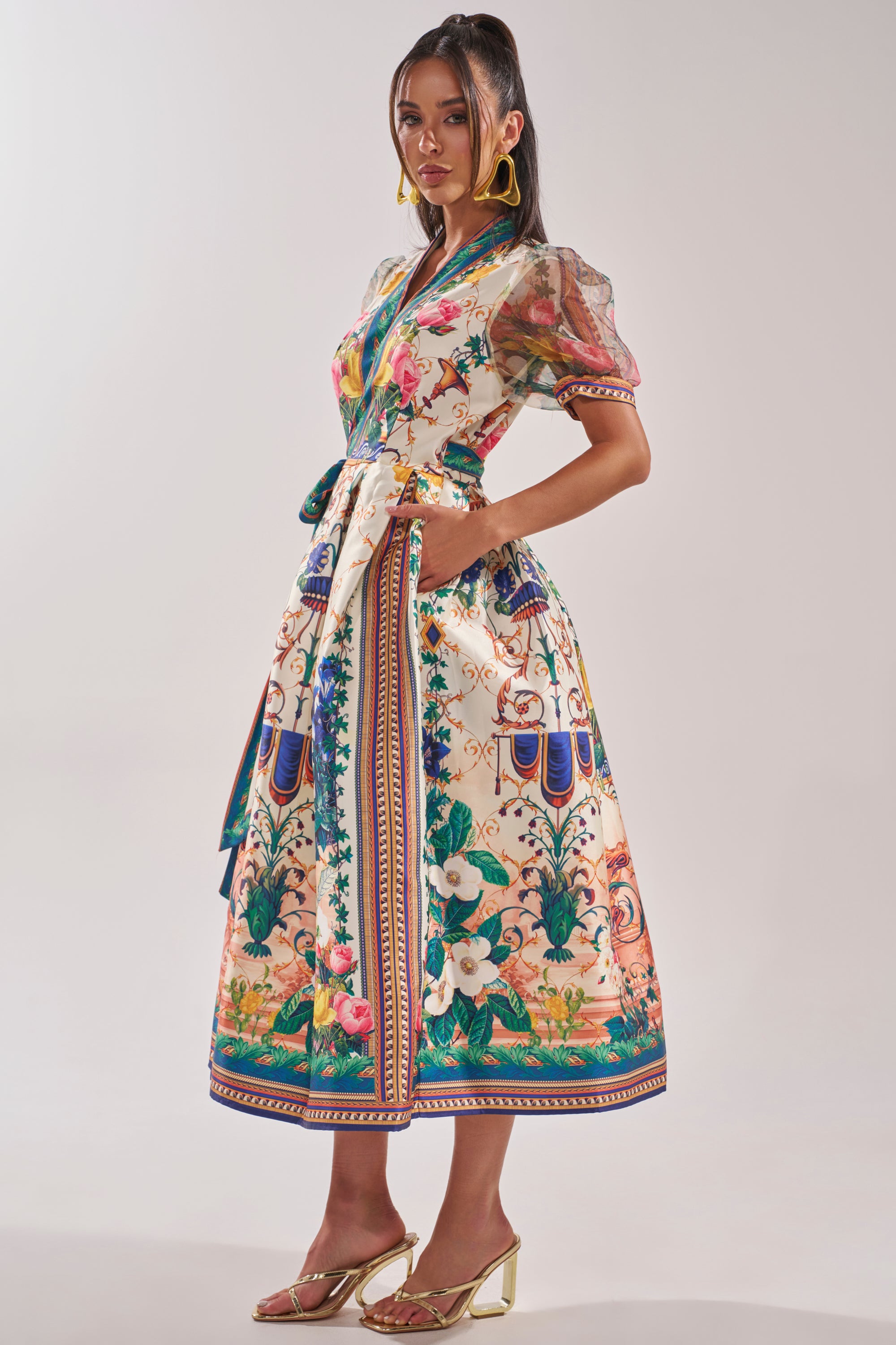 Woman in a FLOWER GIRL SATIN MAXI DRESS featuring botanical print, puff sleeves, and tied waist. She pairs it with gold heels and hoop earrings, has long dark hair, and stands against a plain light background. 3