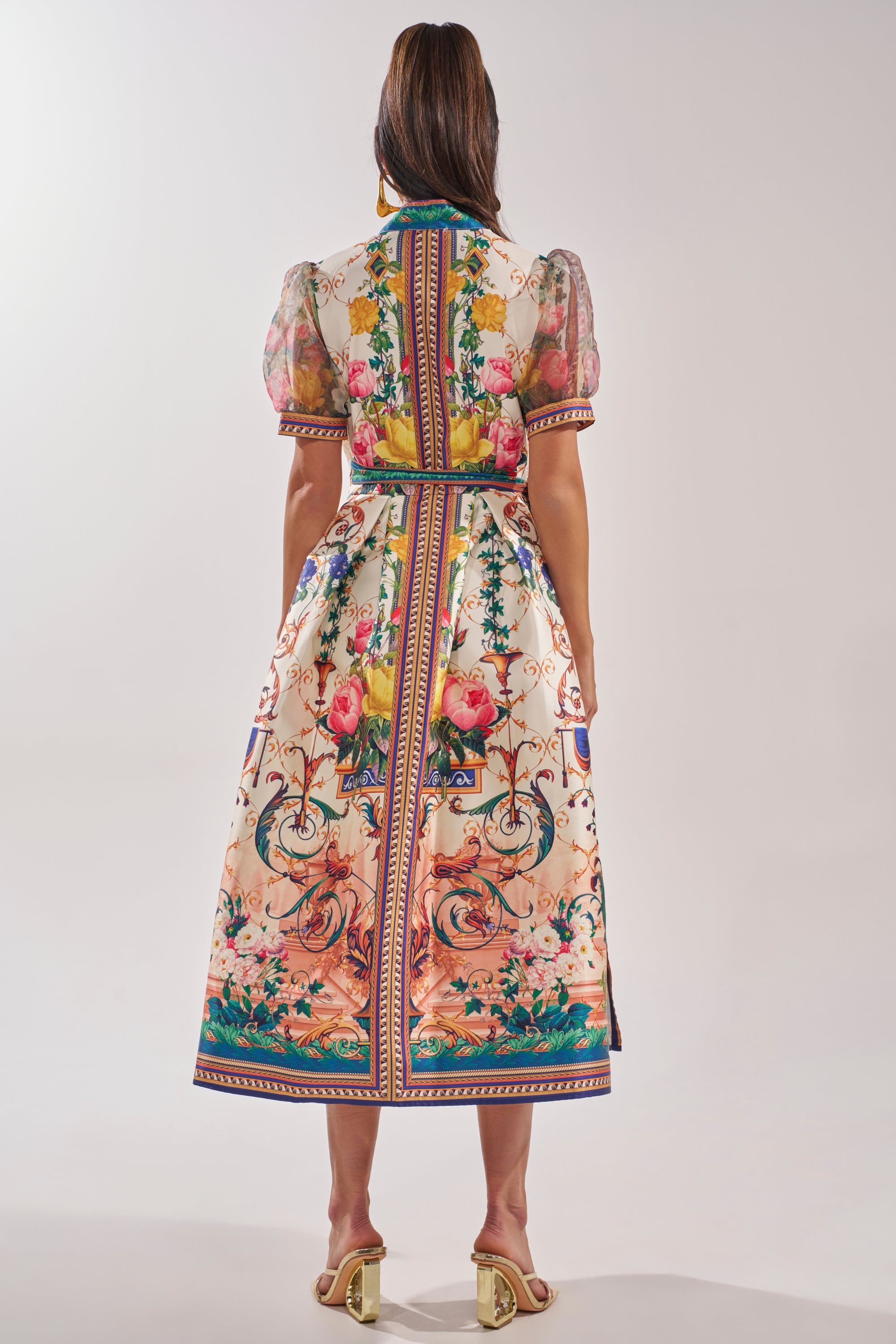A woman faces away, wearing the FLOWER GIRL SATIN MAXI DRESS with puff sleeves and a vibrant botanical print. Her long, straight hair pairs with light-colored open-toe heeled sandals for a chic look. 4