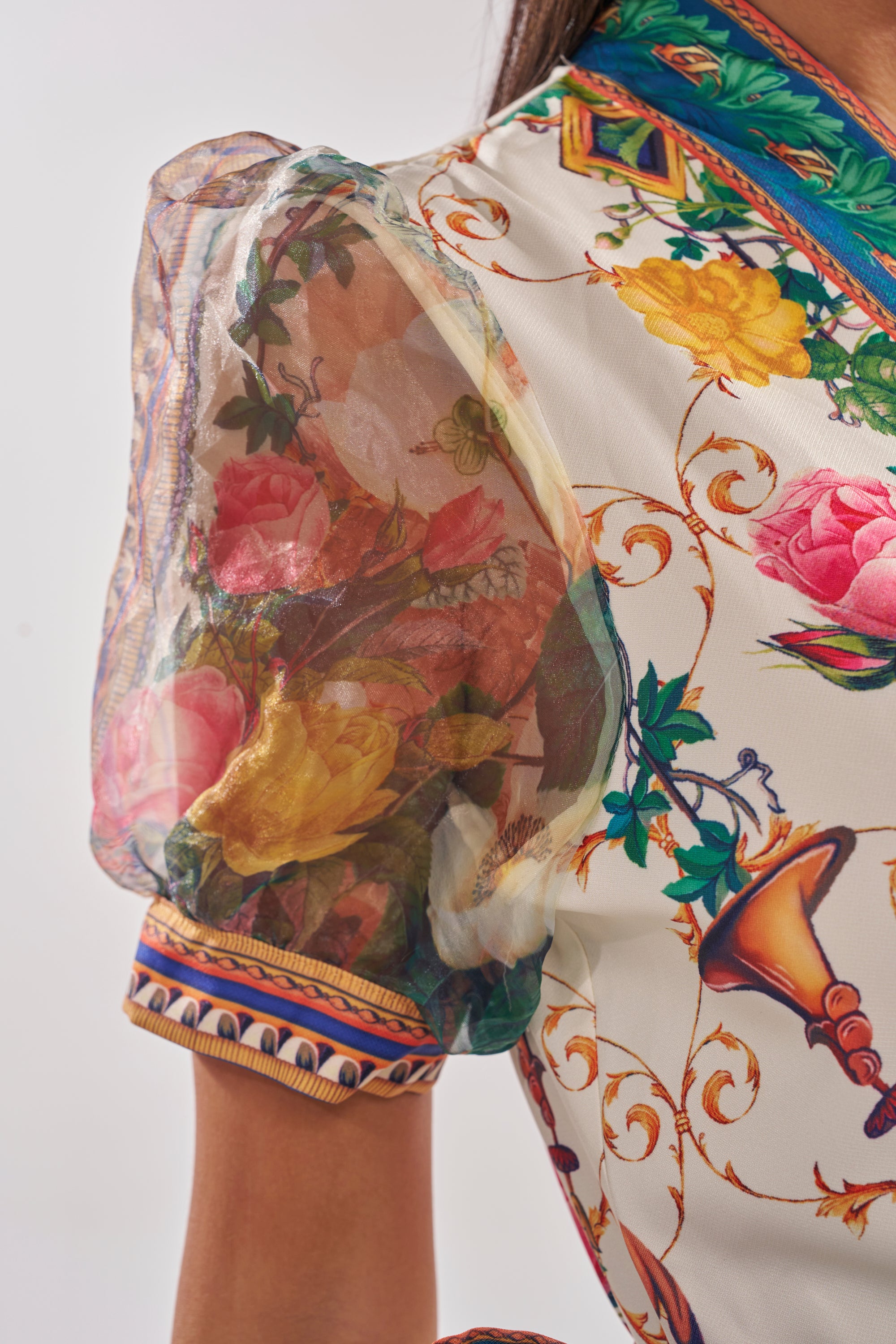 A close-up of a woman's shoulder wearing the FLOWER GIRL SATIN MAXI DRESS, featuring sheer puff sleeves, vibrant multicolored botanical prints, elegant floral patterns, visible stitching, and ornate trim. 5