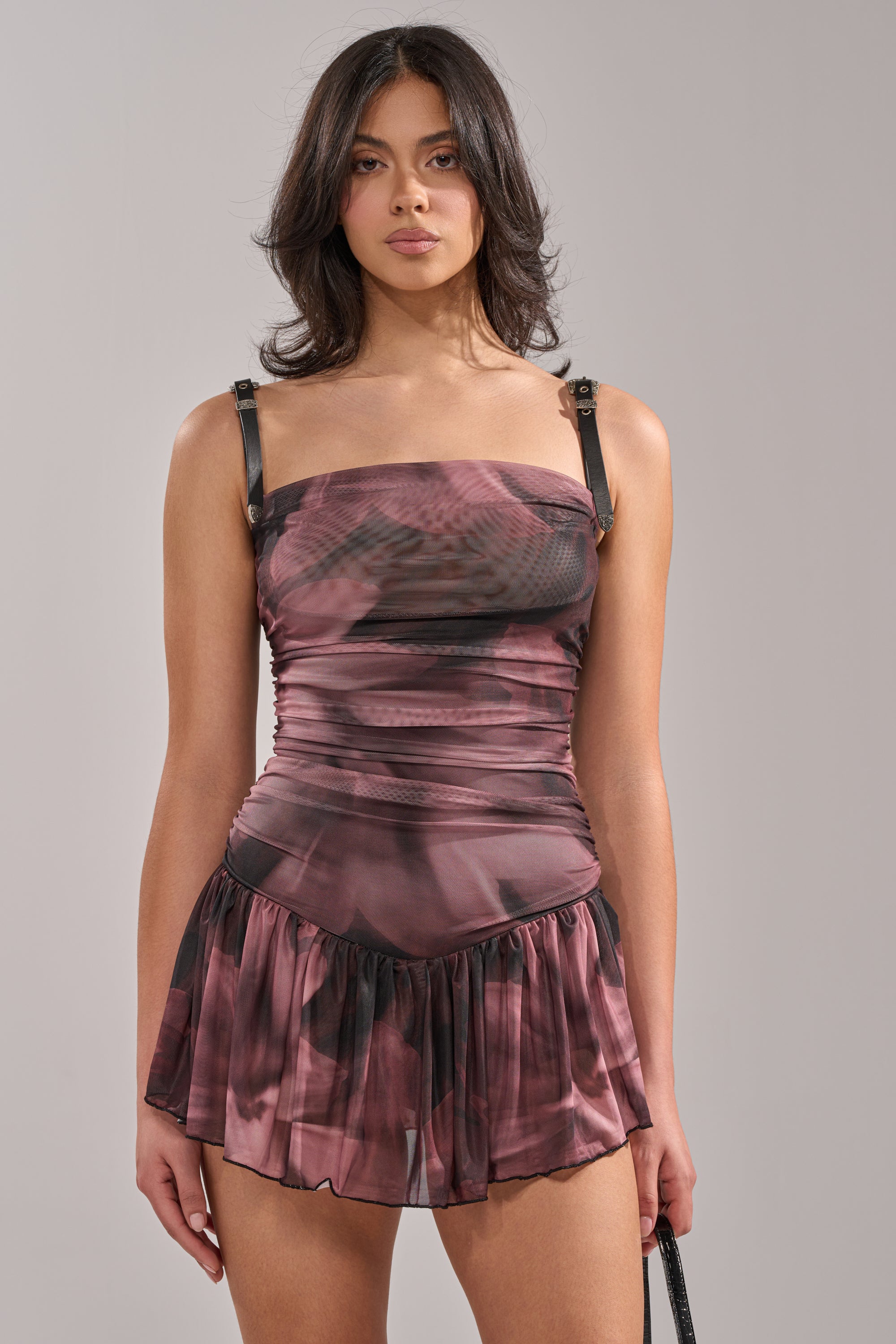 A woman with wavy dark hair wears the JADA MINI DRESS—a sleeveless, stretchy mesh abstract print dress with ruching, thin black straps, and a ruffled skirt—standing against a plain light gray background. 1