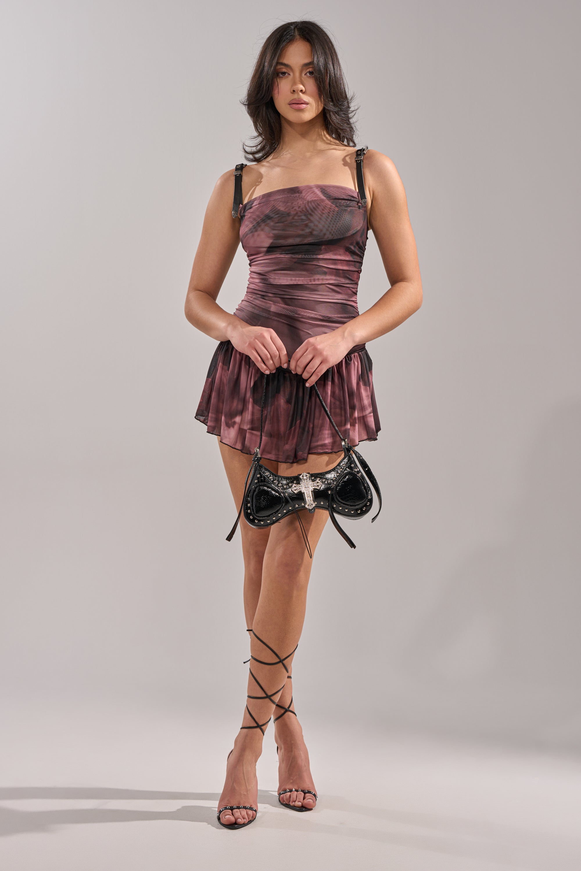 A woman poses against a plain background in the JADA MINI DRESS, an abstract print ruched mini dress with adjustable faux leather straps. She completes the look with lace-up sandals, a black handbag, and loose, wavy hair. 4
