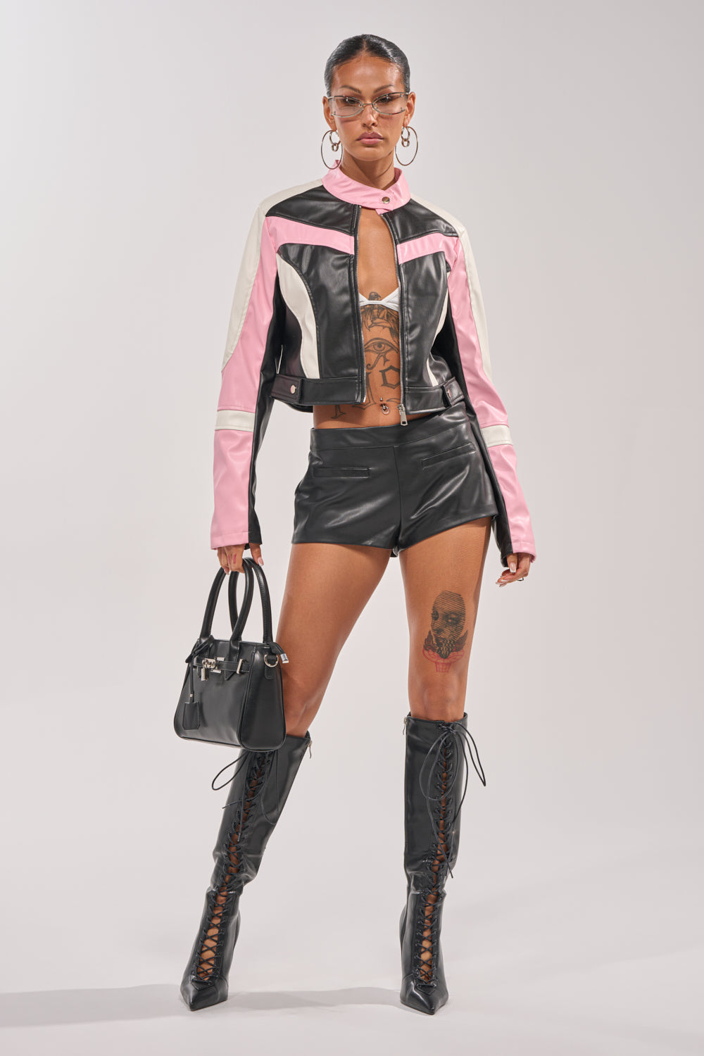 A person styles the LOCKED UP FAUX LEATHER SHORT with a black, pink, and white leather jacket, lace-up knee-high boots, hoop earrings, glasses, and a black handbag, showing tattoos on their chest, stomach, and thigh. 6