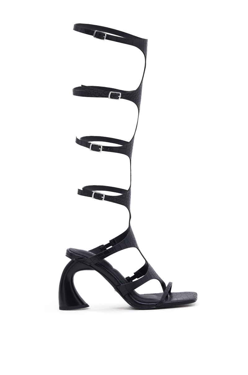 The AZALEA WANG SEAMUS BLACK CROC NOVELTY HEEL SANDAL is a black gladiator-style sandal with five thin buckled straps to the knee, an open toe, and a sculpted chunky heel for a bold, modern statement look.