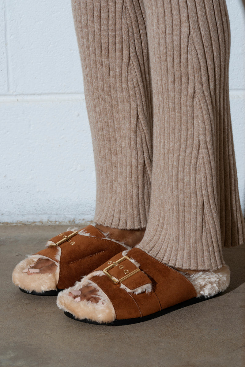 Close-up of a person in beige ribbed knit pants and AZALEA WANG SEANNA TAN FUR SANDAL with a faux suede upper, an open almond toe design, and a faux fur insole, standing on concrete. The white wall behind features a horizontal seam.