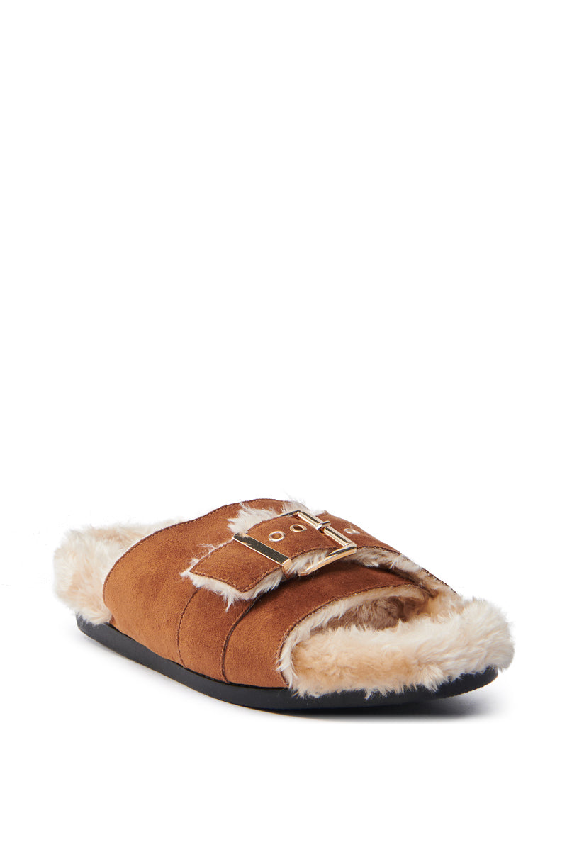 The AZALEA WANG SEANNA TAN FUR SANDAL is a cozy, casual brown slipper with a faux suede upper, faux fur lining, and a gold buckle on top. Featuring an open almond toe and flat black sole, it is set against a white background.