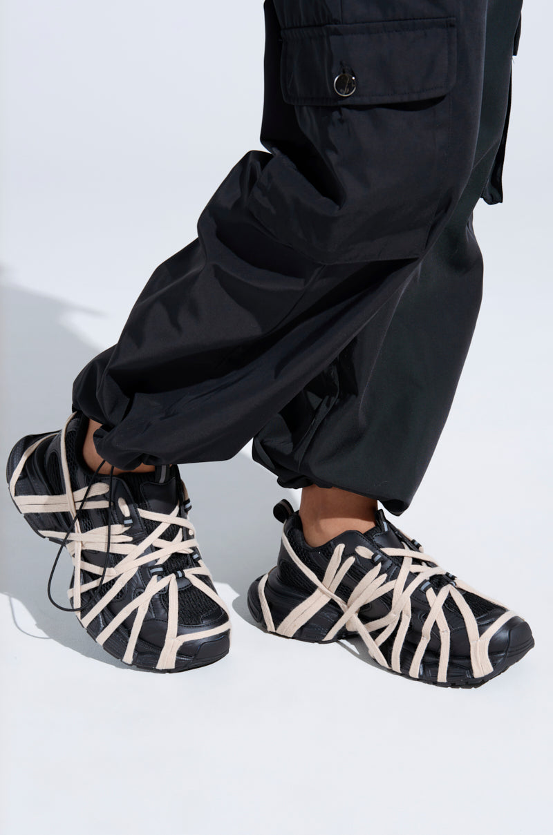 A person in black pants with gathered cuffs and AZALEA WANG SELIE BLACK LACE UP SNEAKERS, featuring thick beige laces and a performance-inspired design, stands on a light surface.