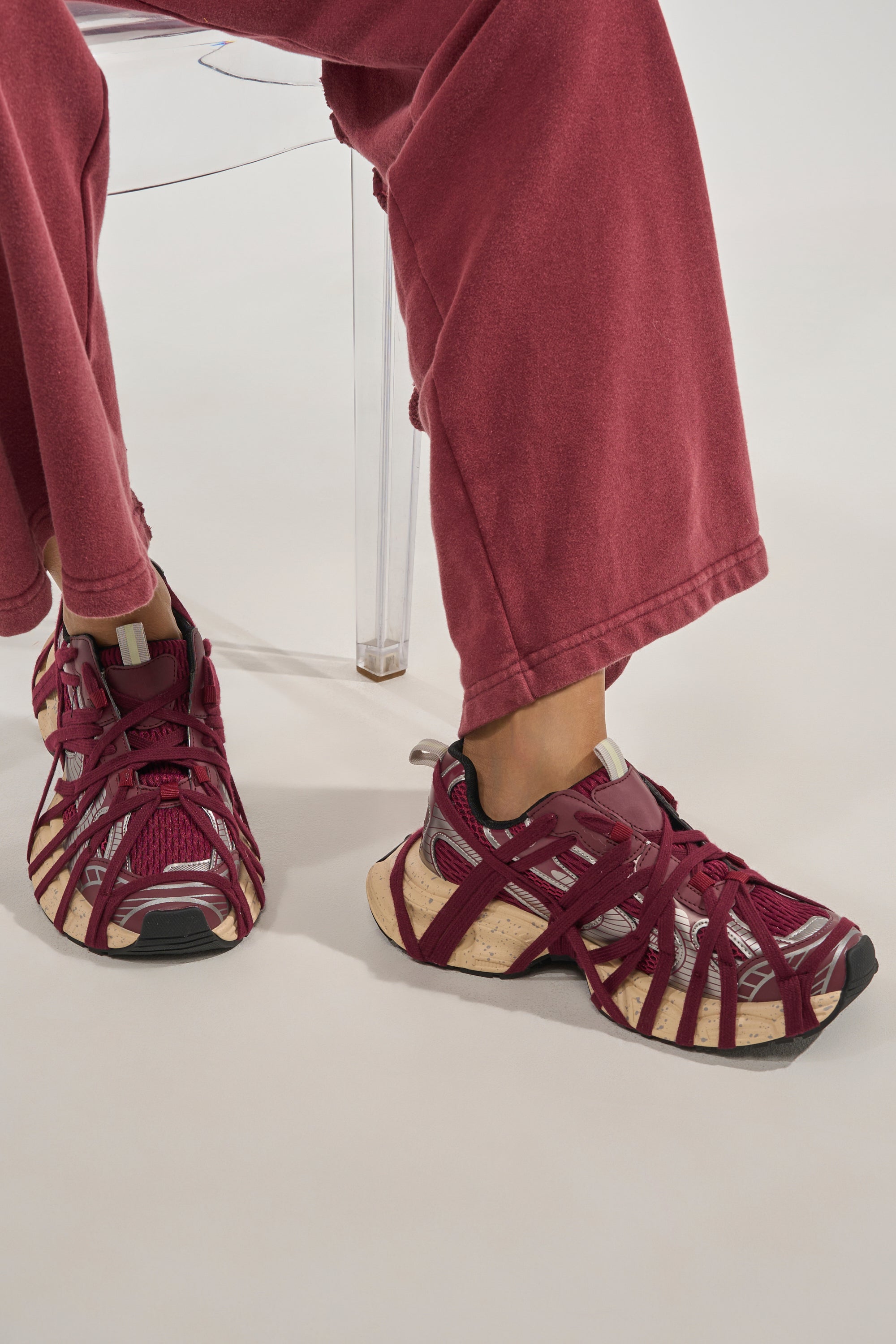 A person in loose red pants and AZALEA WANG SELIE BURGUNDY LACE UP SNEAKER sits on a clear acrylic chair, their feet resting on a white surface. 0