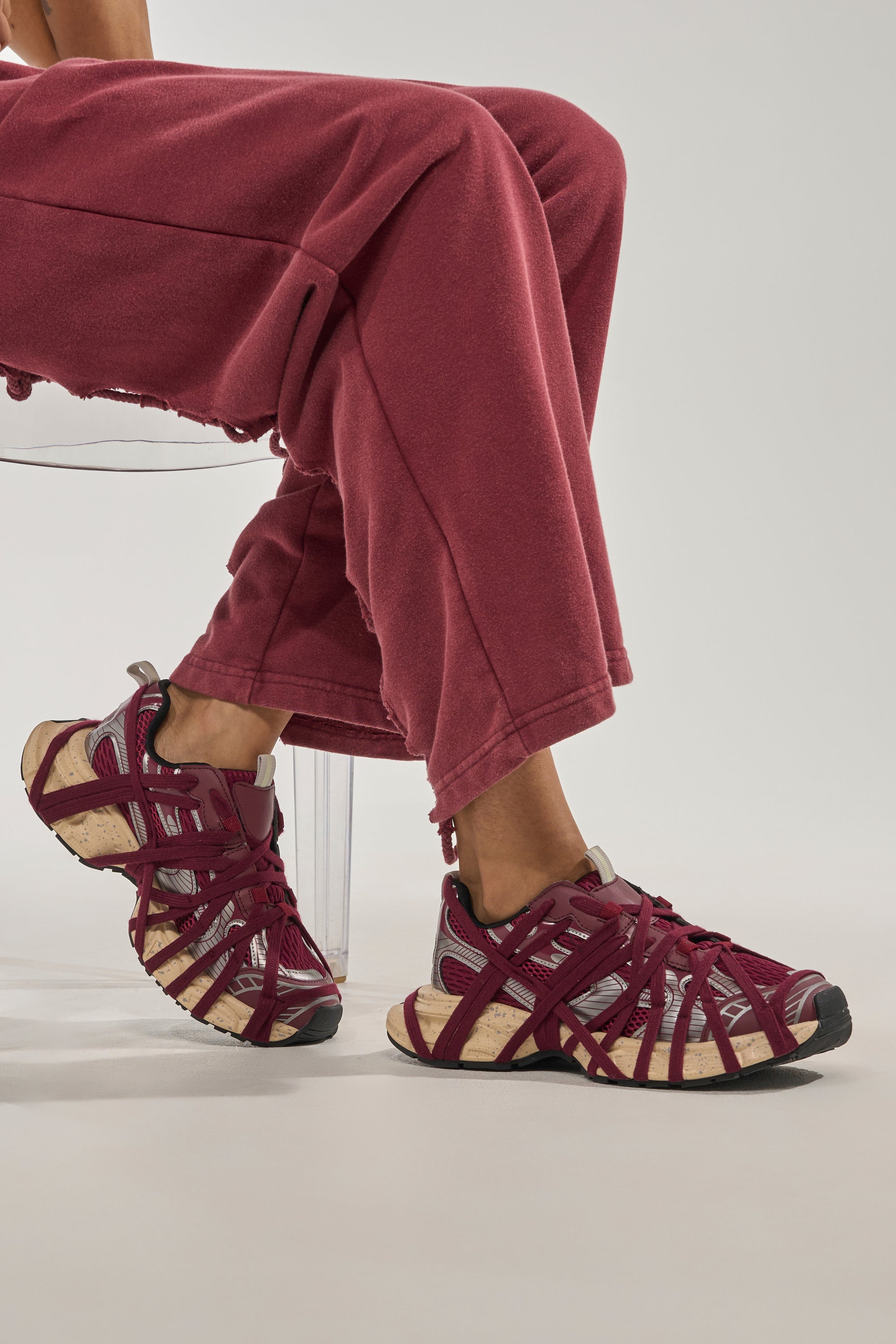 A person in loose maroon pants sits on a clear chair, highlighting the AZALEA WANG SELIE BURGUNDY LACE UP SNEAKER with bold crisscrossing straps. The plain background makes these sporty, chunky sneakers stand out. 1