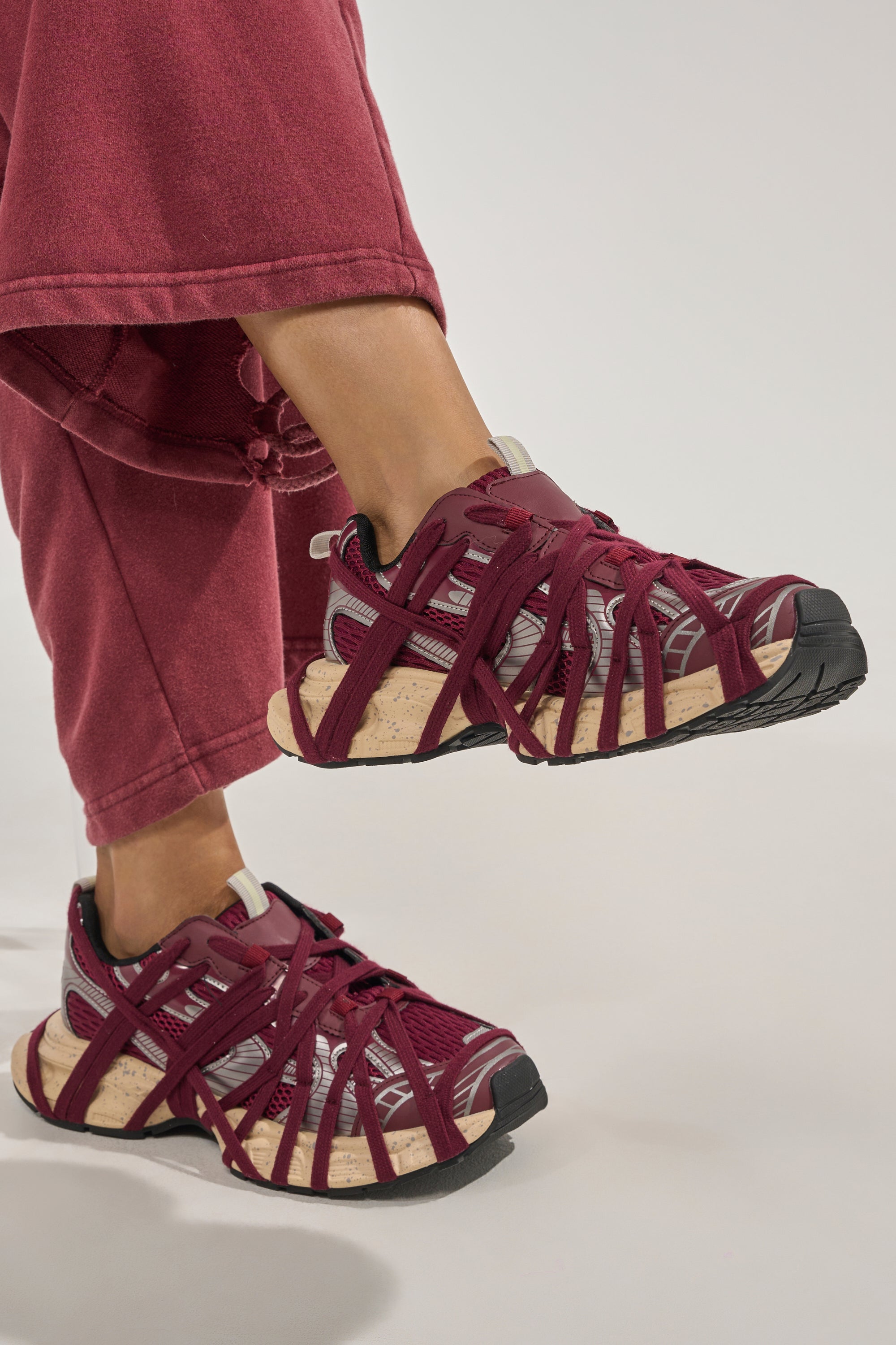 Wearing AZALEA WANG SELIE BURGUNDY LACE UP SNEAKERS with crisscross straps and beige soles, a person in matching burgundy pants lifts one foot slightly off the ground against a plain background. 6