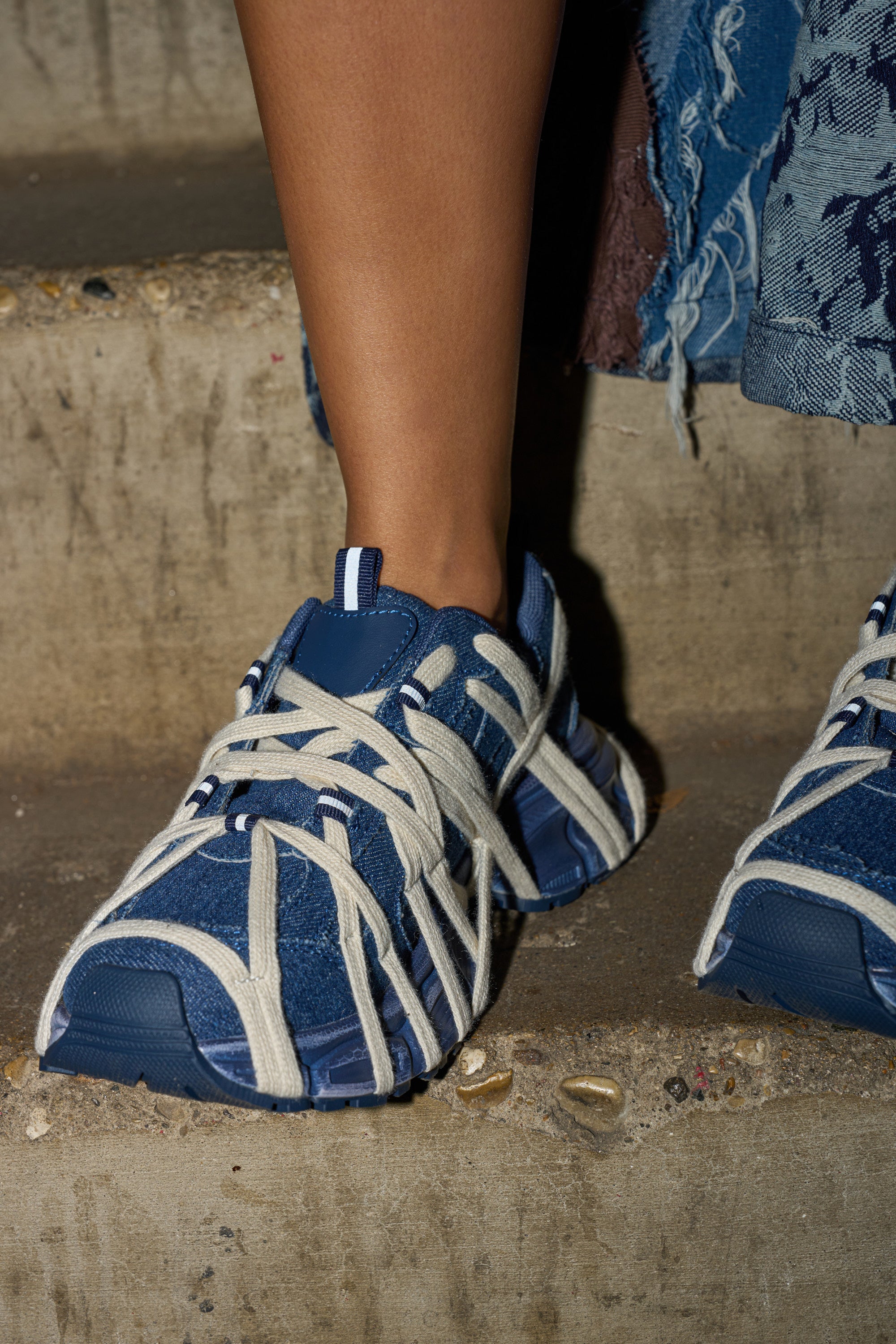 Someone wearing AZALEA WANG SELIE DENIM LACE DETAIL SNEAKER stands on concrete steps, with part of a frayed blue denim skirt visible.
