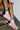 A person sits on the stairs, sporting striped socks and colorful AZALEA WANG SELIE RED FLAT SNEAKERS with a unique textile upper. The backdrop showcases a textured metal step against a white brick wall, highlighting the playful footwear design.