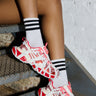 A person sits on the stairs, sporting striped socks and colorful AZALEA WANG SELIE RED FLAT SNEAKERS with a unique textile upper. The backdrop showcases a textured metal step against a white brick wall, highlighting the playful footwear design.