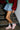 A person wearing a red fur jacket, green shirt, denim skirt, and AZALEA WANG SELIE RED FLAT SNEAKER with a textile upper and platform sole stands on metal stairs against a white brick wall.
