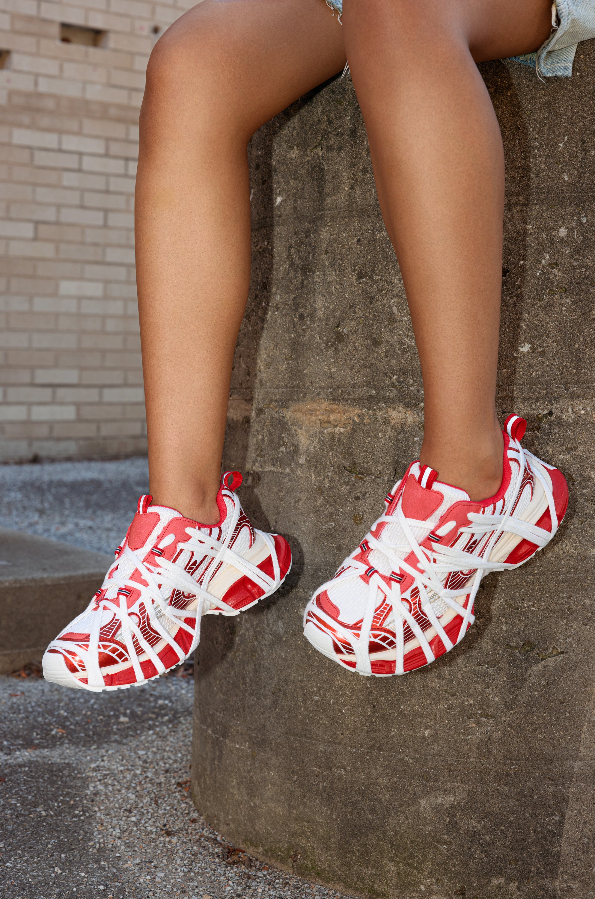 A person wearing AZALEA WANG SELIE RED FLAT SNEAKER sits on a concrete ledge with legs extended and feet dangling, set against a brick wall and gravel ground for an urban vibe.