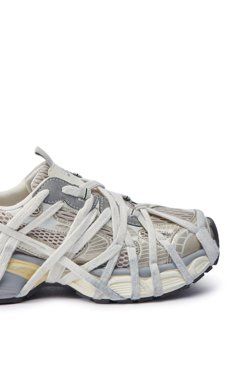 Close-up of the AZALEA WANG SELIE WHITE FLAT SNEAKER, showcasing a mesh upper in gray and beige tones. A web-like design of multiple overlapping white laces enhances its style, while the slip-on sneaker is paired with a thick, textured rubber sole featuring multi-tone sections for a modern look.