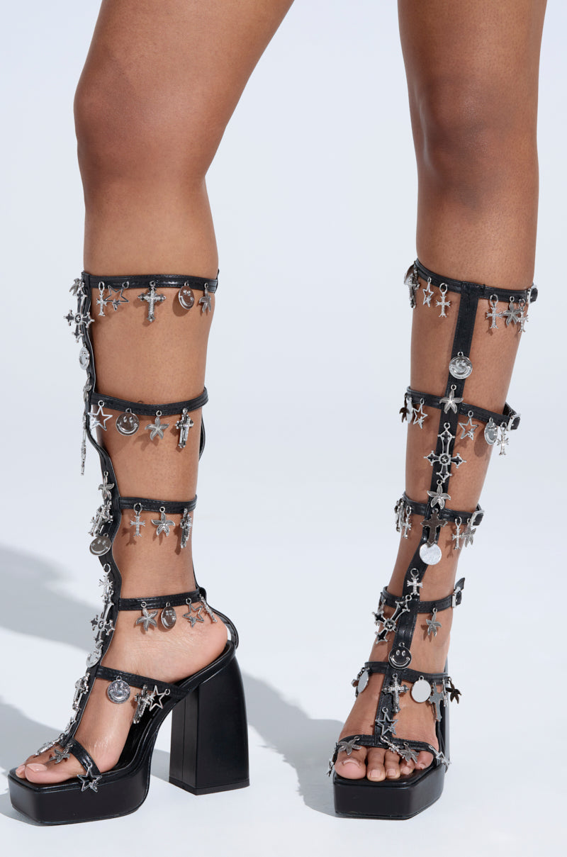 Close-up of a person wearing the AZALEA WANG SELINDE BLACK SILVER METAL HARDWARE CHUNKY HEEL SANDAL, featuring high-heeled, open-toe gladiator design with platform soles. The black faux leather straps are adorned with silver charms like crosses and circles, extending just below the knee.