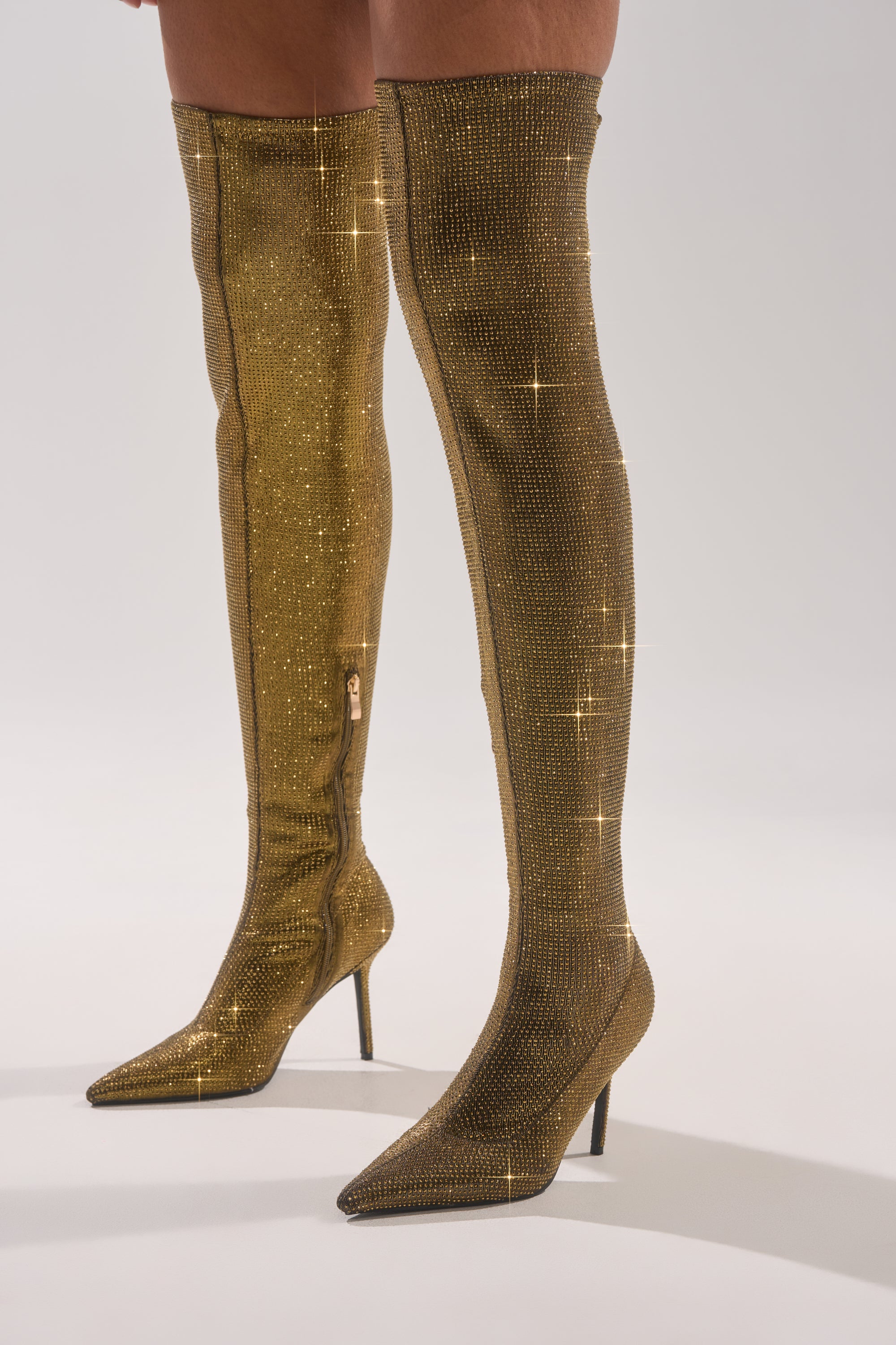 A person wears the AKIRA GOLD SELMA GOLD SPARKLE OVER THE KNEE BOOT, featuring stiletto heels, pointed toes, and glittery gold material that reflects light for a shiny, glamorous look.