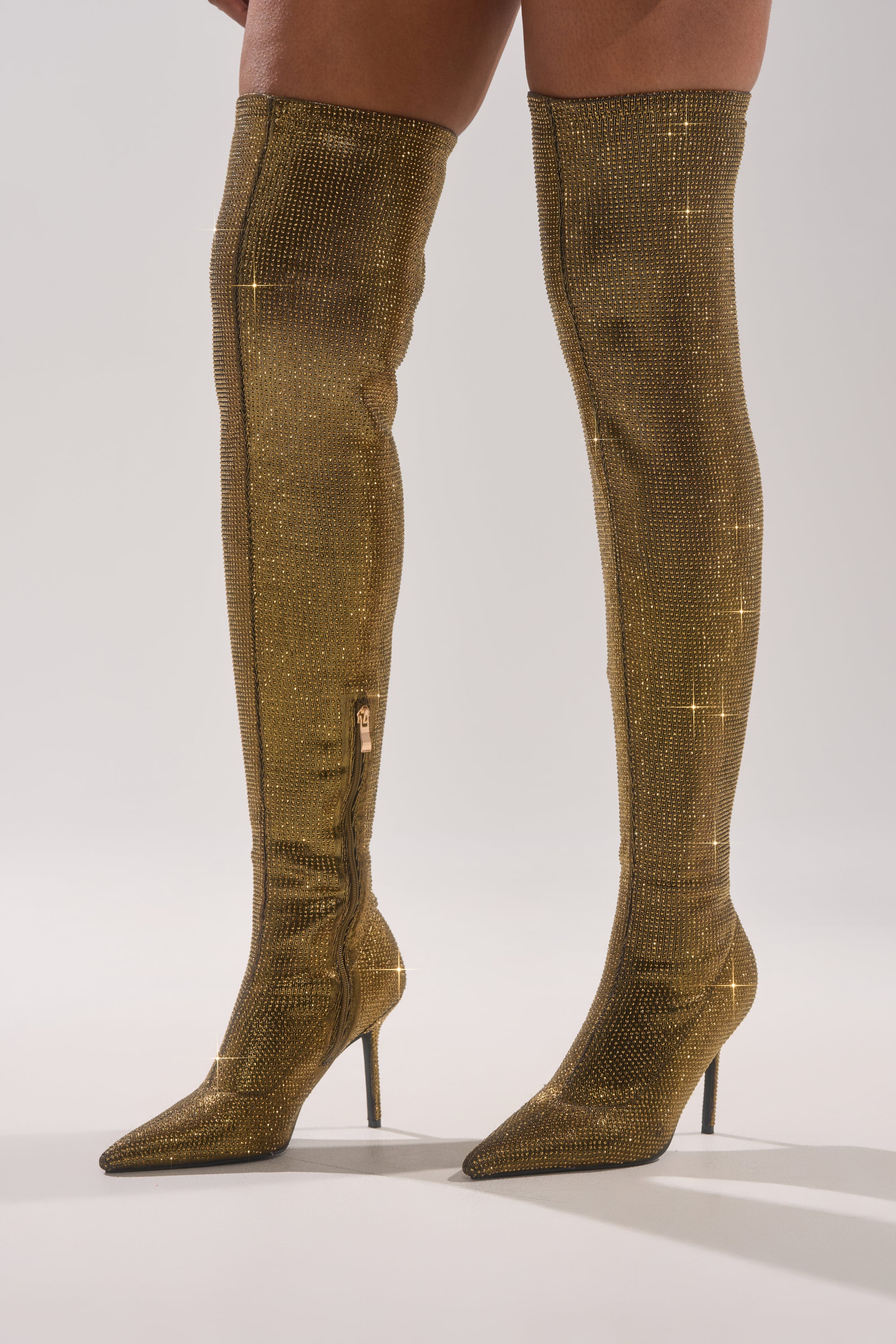 A person models the AKIRA GOLD SELMA GOLD SPARKLE OVER THE KNEE BOOT—rhinestone-covered thigh-high boots with pointed toes and stiletto heels—against a plain light background.