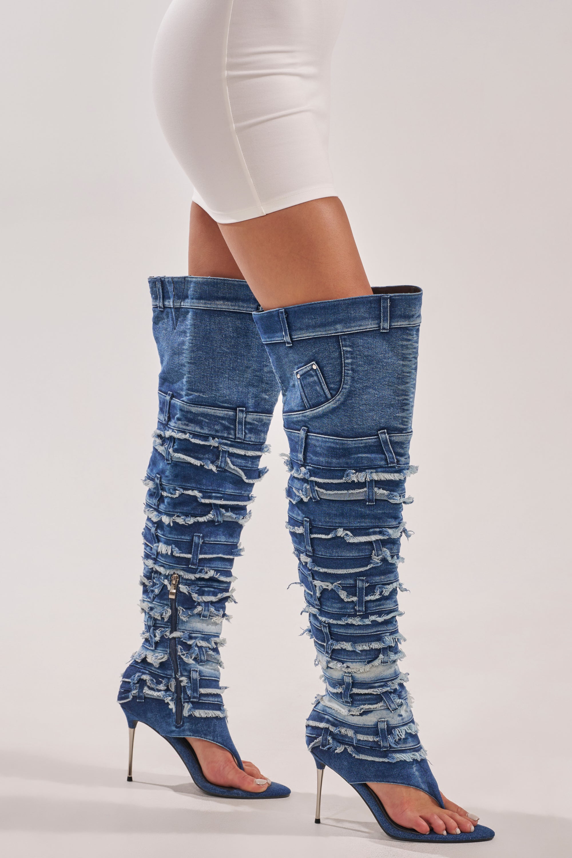 A person stands against a plain light background, wearing a white fitted skirt and the AZALEA WANG SENA DENIM STILETTO OPEN TOE BOOT—distressed, thigh-high denim boots with frayed patches and sleek stiletto heels. 0