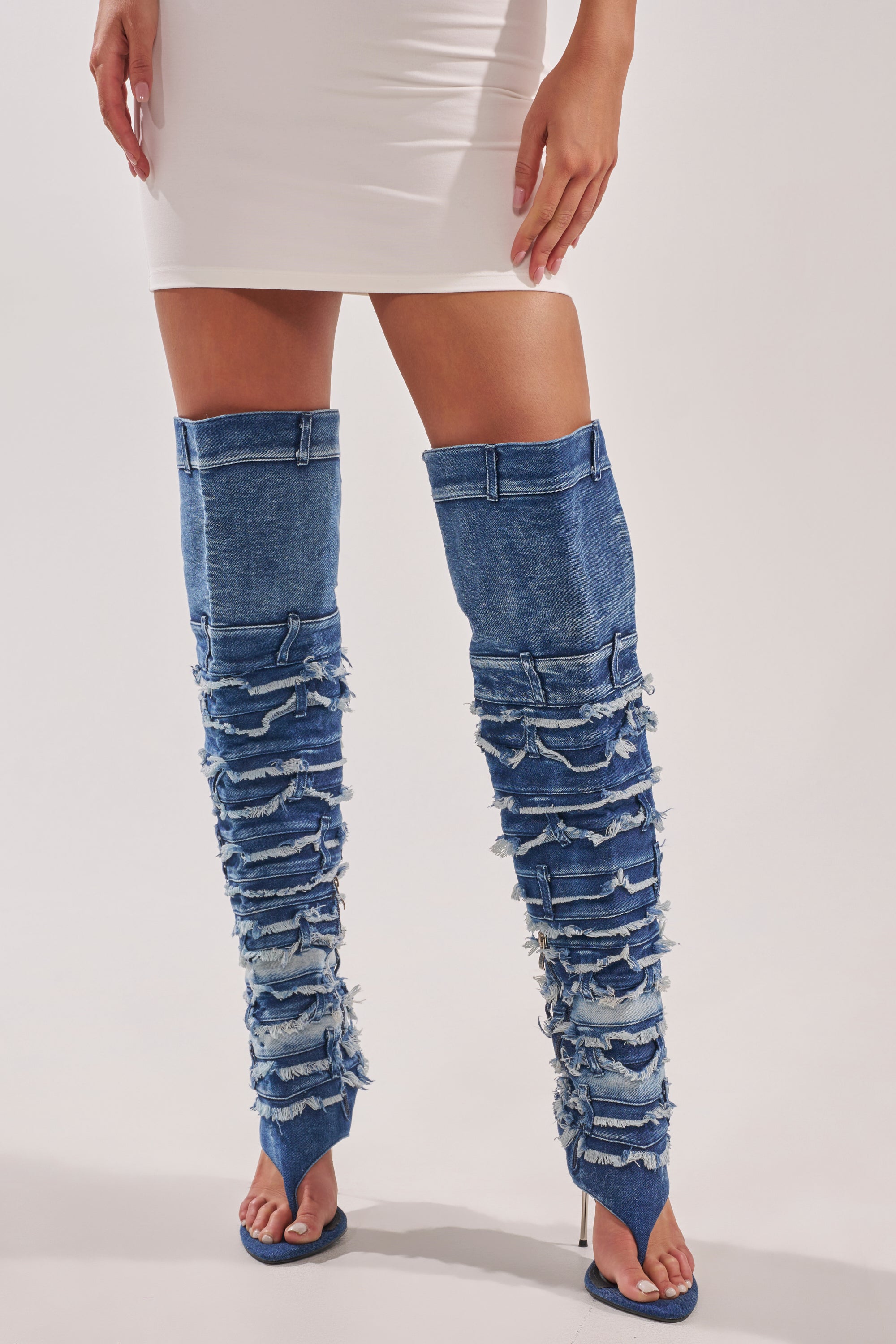 A person in a white mini dress and AZALEA WANG SENA DENIM STILETTO OPEN TOE BOOTS with frayed details and high heels stands against a plain background. 1