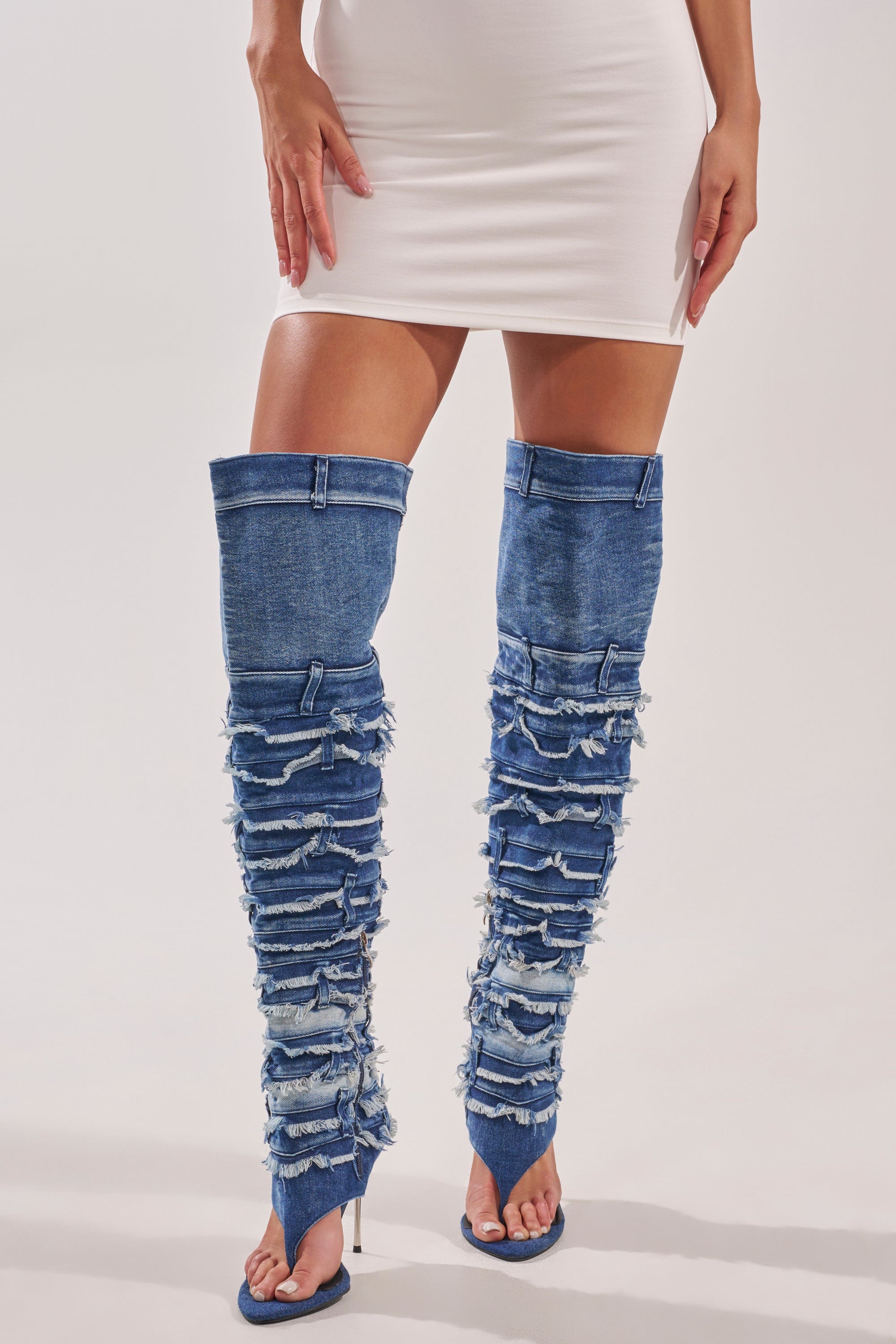 A person wearing a white mini dress and the AZALEA WANG SENA DENIM STILETTO OPEN TOE BOOT, featuring distressed thigh-high denim, open toes, and high heels, stands against a plain background. 2
