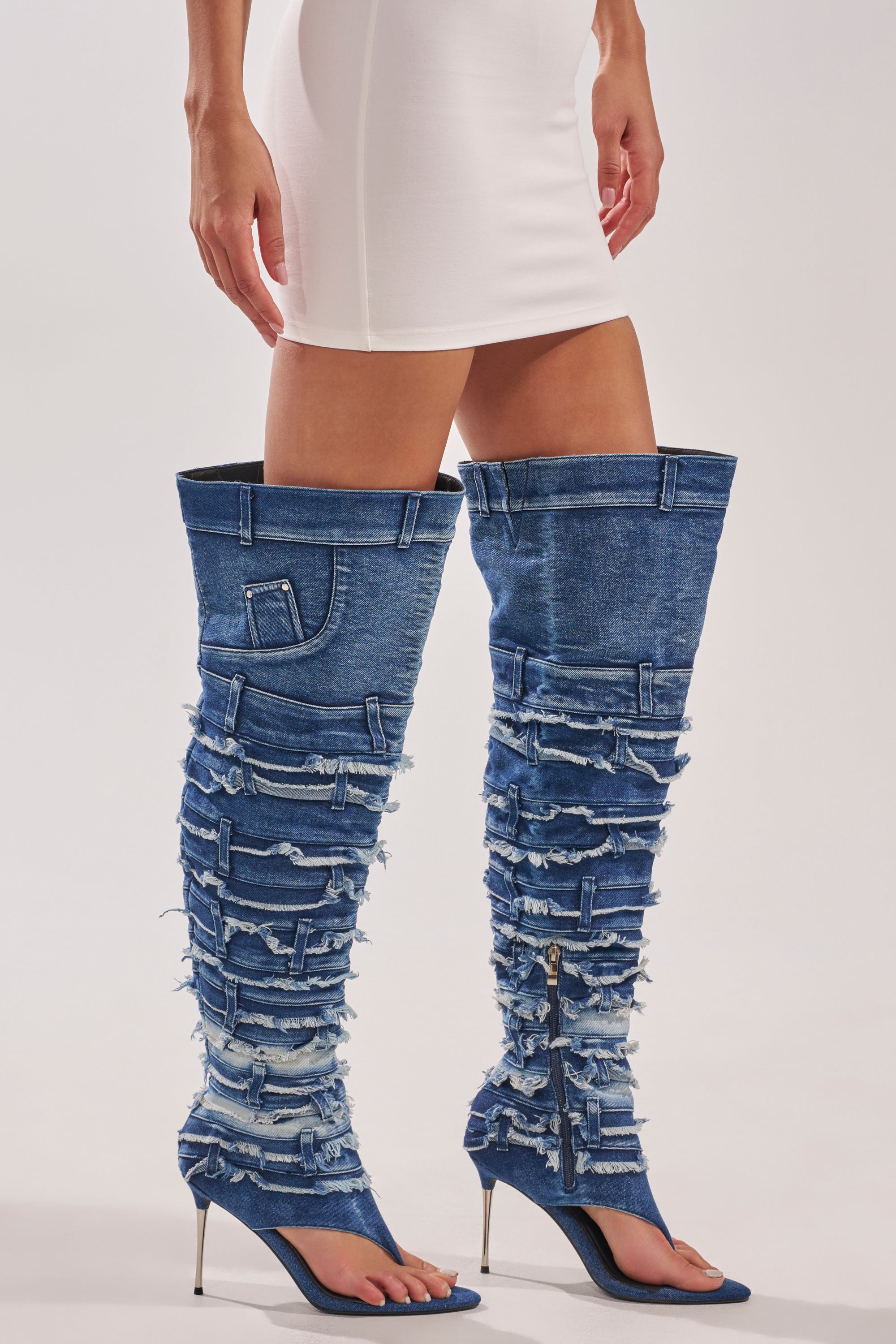 A person wearing a white mini dress and bold AZALEA WANG SENA DENIM STILETTO OPEN TOE BOOTS featuring unique ripped details, pockets, zippers, and a striking stiletto heel. 3