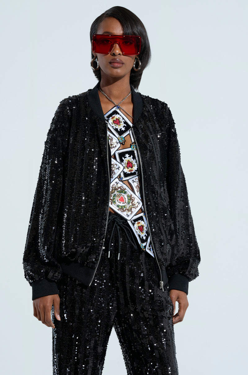A person wearing red sunglasses and a COSMIC SEQUIN BOMBER adorned with glittering sequin embellishments, paired with matching pants, is seen. Underneath, they wear a shirt with a geometric pattern while standing against a light-colored background. 0