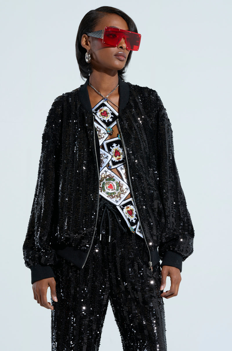 A person sporting vivid red sunglasses and the striking COSMIC SEQUIN BOMBER, adorned with shimmering sequin details, paired with a patterned top featuring lively designs. They have short black hair and are positioned against a simple light backdrop. 1
