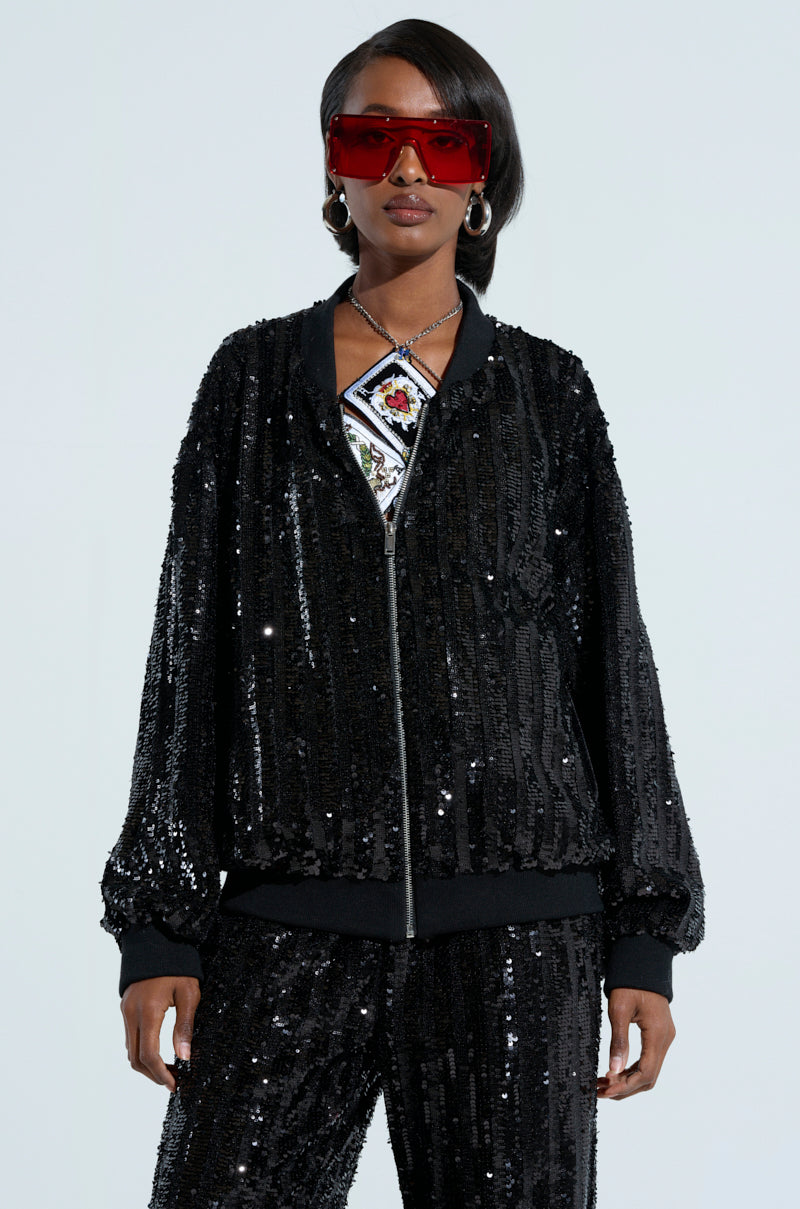 A person dressed in a COSMIC SEQUIN BOMBER and pants, complemented by large red sunglasses, hoop earrings, and a pendant necklace. The outfit's dazzling sequin embellishments catch the light, generating a shimmering effect against the minimal background. 2