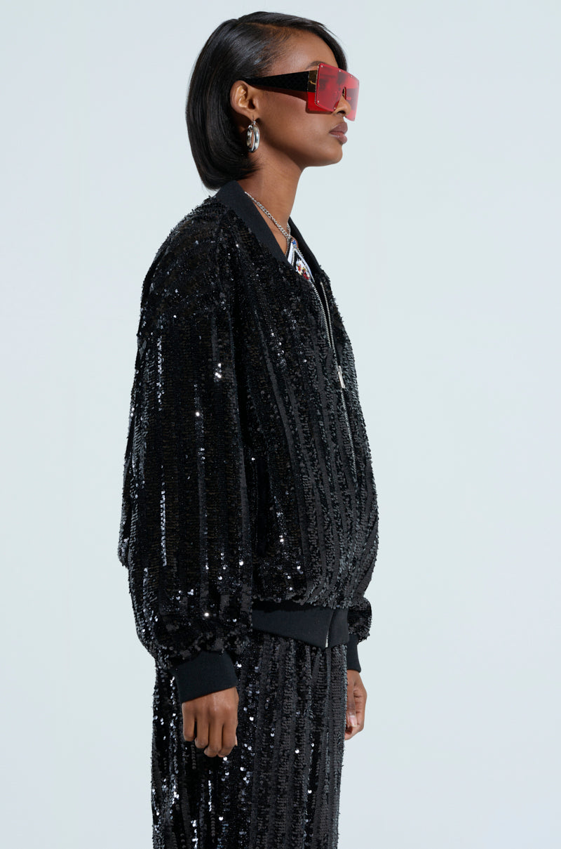 A person with short black hair is seen in a side profile view, sporting oversized red sunglasses and the COSMIC SEQUIN BOMBER, which dazzles with glittering sequin embellishments. The extraordinary outfit is paired with matching pants and set against a light gray background. 3