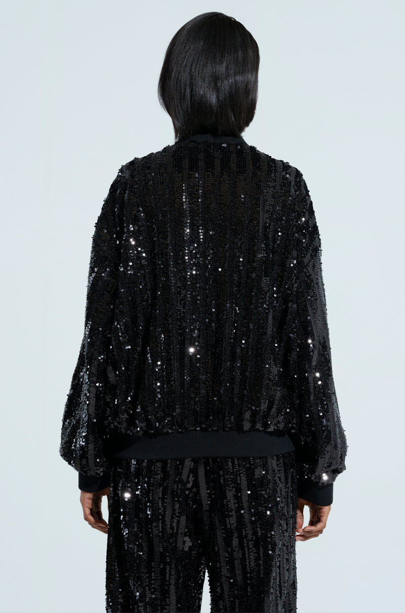 A person with long dark hair stands facing away, dressed in a COSMIC SEQUIN BOMBER that sparkles. Both the jacket and pants are adorned with glittering sequin embellishments, subtly reflecting light against a plain background. 4