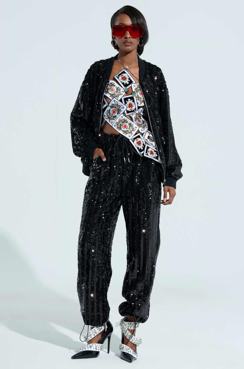 A person confidently stands in a dazzling black COSMIC SEQUIN BOMBER and colorful embroidered top. They pair this striking outfit with red sunglasses and chic black-and-white heeled shoes, all set against a light-colored, plain background that perfectly enhances their vibrant style. 6