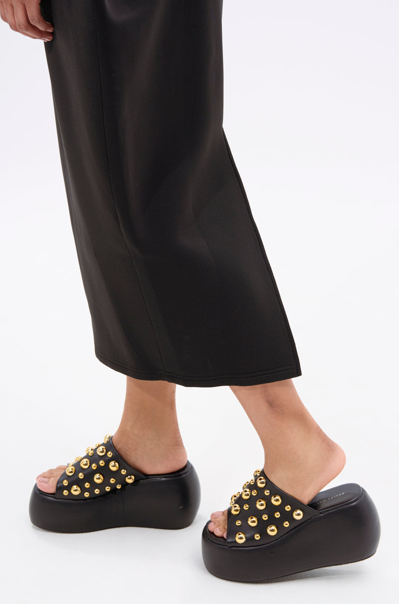 A person in black wide-leg pants stands on a white surface, wearing the AZALEA WANG SERGO BLACK EMBELLISHED CHUNKY SANDAL with a faux leather platform sole.