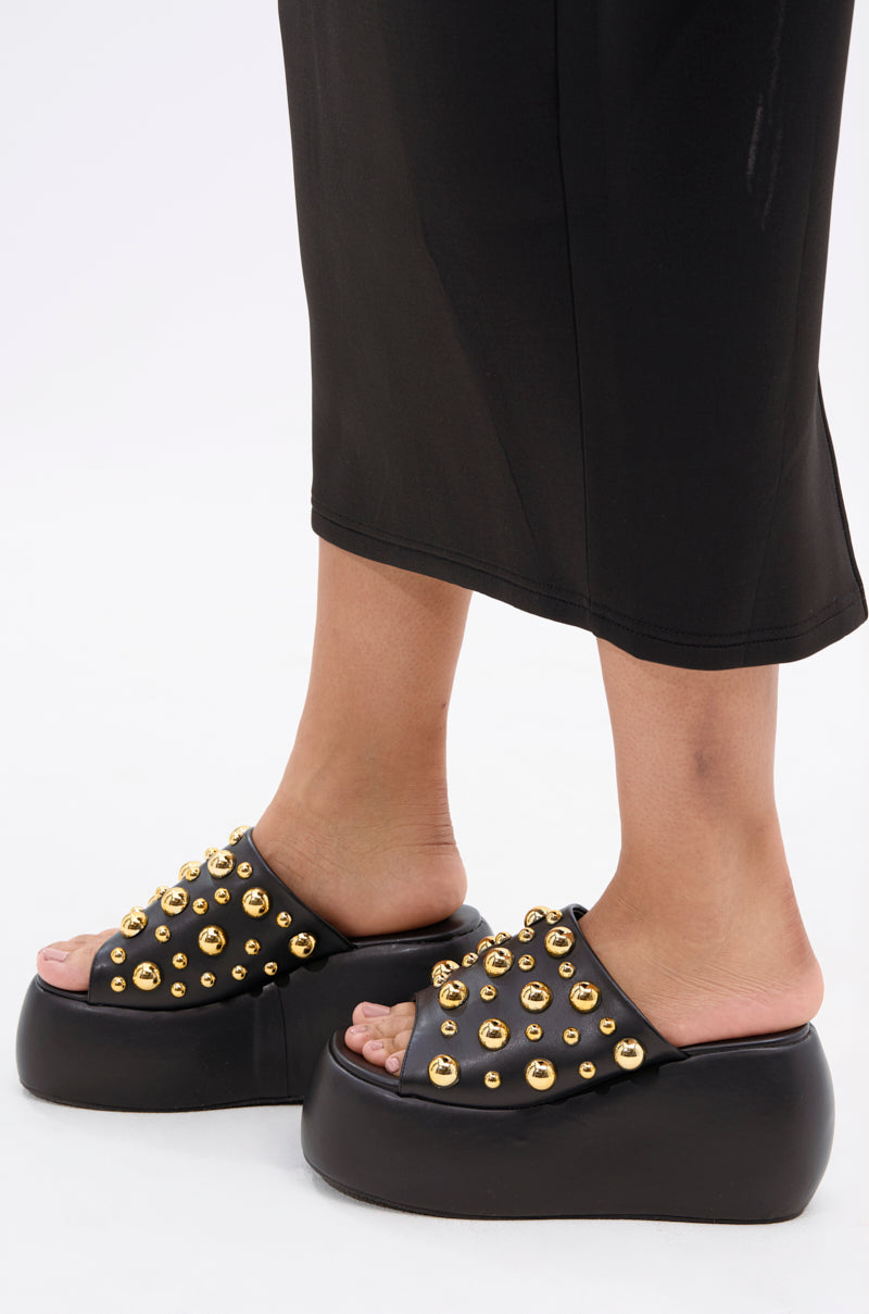 A person in a black midi skirt and AZALEA WANG SERGO BLACK EMBELLISHED CHUNKY SANDAL with a platform sole and multiple gold studs stands on a white background.