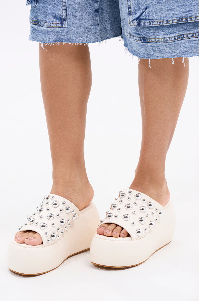 The AZALEA WANG SERGO BONE EMBELLISHED CHUNKY SANDAL, featuring silver studs and a platform heel, is styled with light denim shorts. Photo focuses on legs and feet against a white backdrop, highlighting the sandal's bold details.