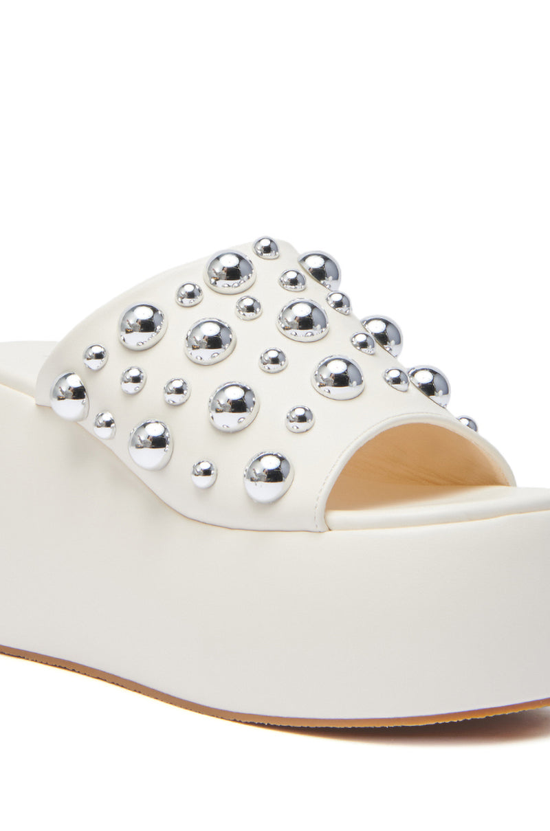 The AZALEA WANG SERGO BONE EMBELLISHED CHUNKY SANDAL is a white faux leather platform sandal with an open toe, chunky heel, and a wide strap adorned with shiny metallic studs in varying sizes.