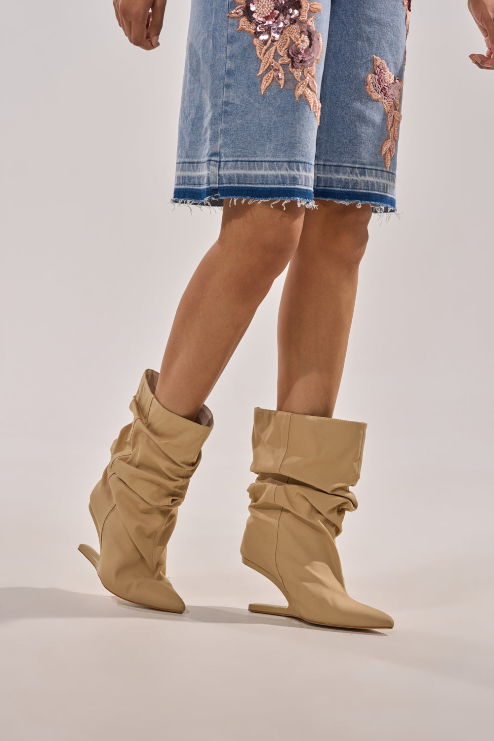 A person wearing the AZALEA WANG SERONA KHAKI INVISIBLE WEDGE BOOTIE and a knee-length denim skirt with floral embroidery and a frayed hem stands against a plain background. 0