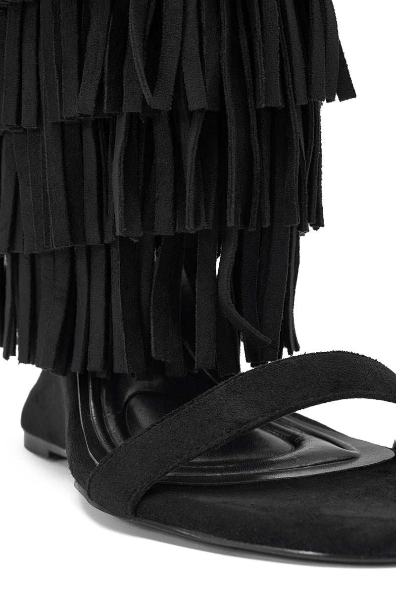 A close-up of the AKIRA GOLD SESTINA BLACK FAUX SUEDE FRINGE WEDGE SANDAL shows its thick strap, suede-like texture, and black fringe draping over the foot for a chic, relaxed mid-calf look. 6