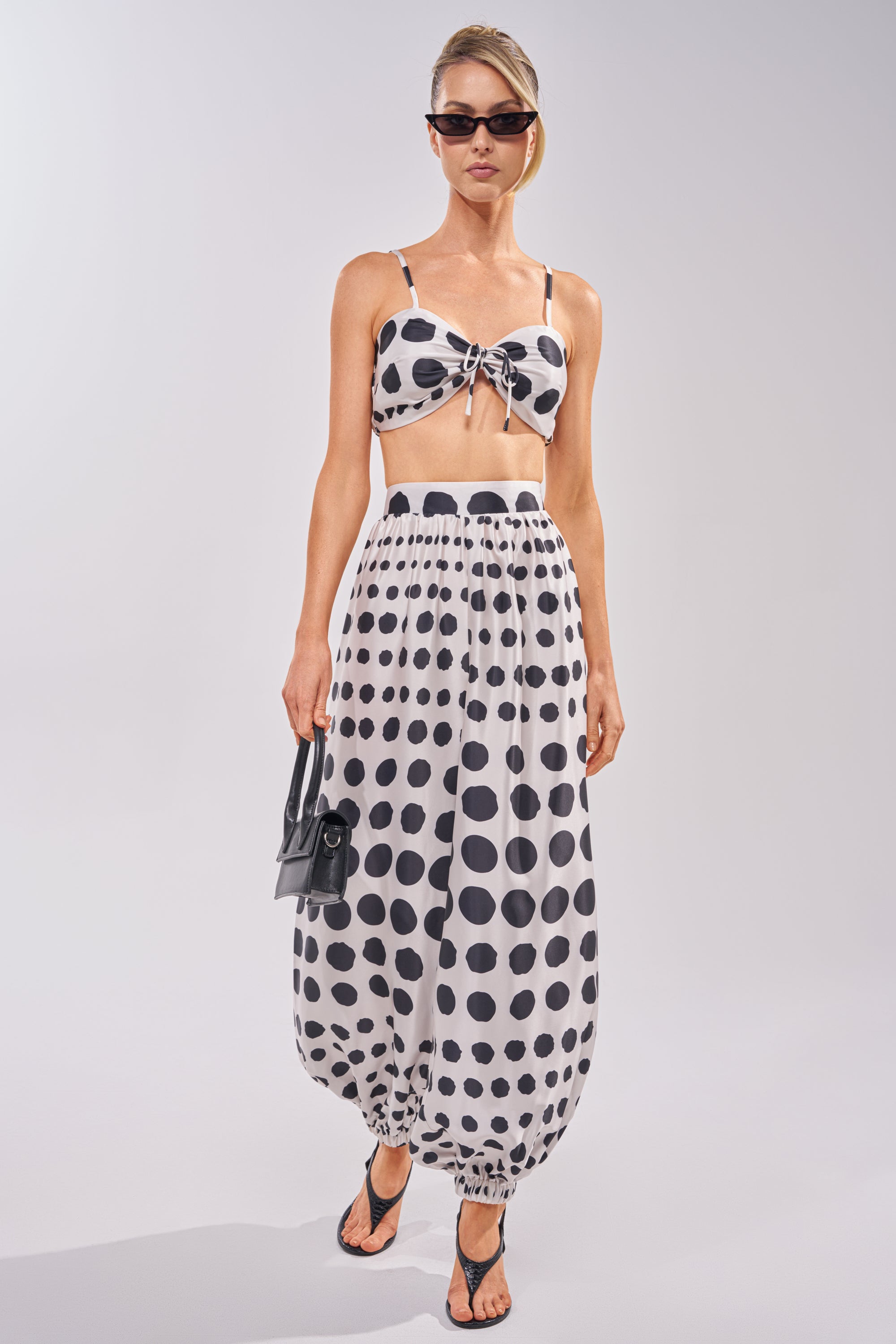 A woman wears the POLKA DOT BIKINI GIRL SET—sunglasses, a black and white polka dot bralette with matching high-waisted balloon pants—paired with black sandals and a small black handbag. 1