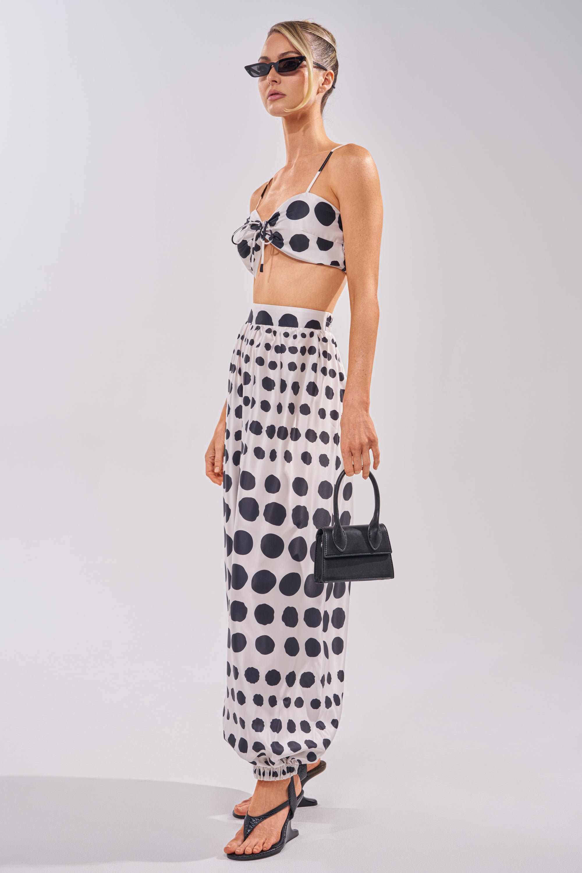 A woman wearing sunglasses and the POLKA DOT BIKINI GIRL SET—a matching white bralette and long skirt with large black polka dots—black sandals, and a small black handbag stands against a plain background. 2