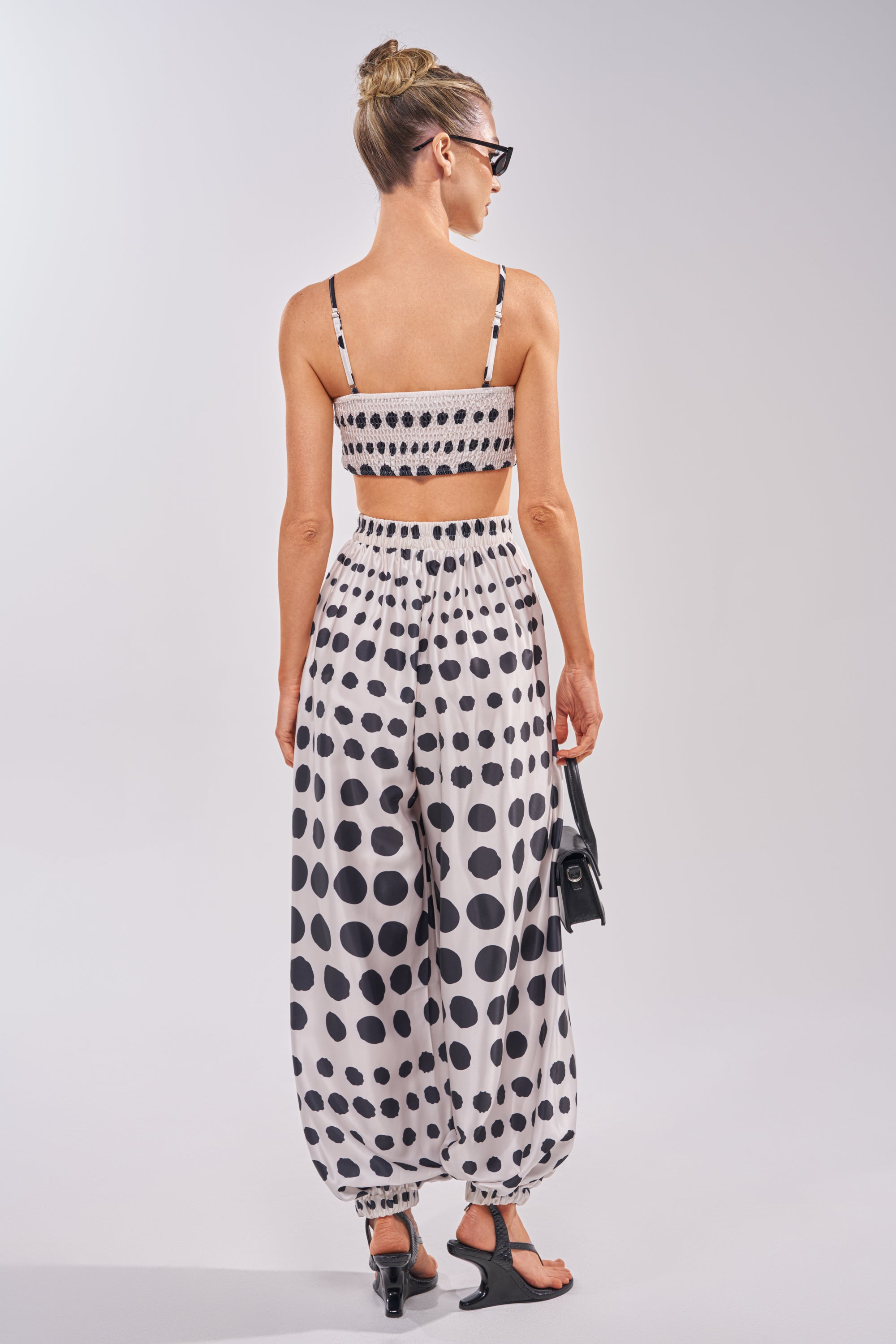 A woman with blonde hair in a bun and sunglasses wears the POLKA DOT BIKINI GIRL SET with matching high-waisted pants, black sandals, and a small black handbag, facing away from the camera. 3