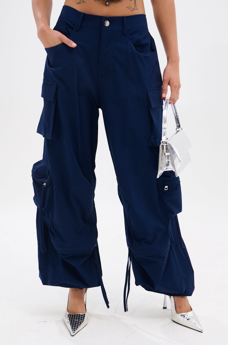 A person wearing FEEL GOOD TROUSER cargo pants in navy blue with a high-rise elastic waistband and cargo pockets, paired with a small silver handbag and pointed mesh-detailed silver heels.