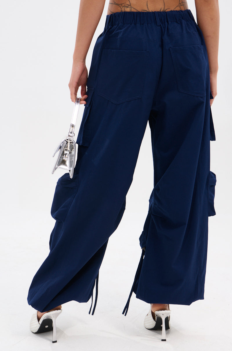 A person is shown from the back wearing FEEL GOOD TROUSER navy wide-leg cargo pants with a high-rise elastic waistband and cargo pockets, paired with white high-heeled shoes and holding a small transparent handbag.
