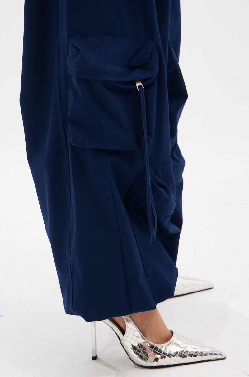 A person stands on a white surface wearing the FEEL GOOD TROUSER in navy, featuring wide legs, cargo pockets, and a high-rise elastic waistband, paired with pointed silver geometric-patterned heels.