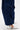A person stands on a white surface wearing the FEEL GOOD TROUSER in navy, featuring wide legs, cargo pockets, and a high-rise elastic waistband, paired with pointed silver geometric-patterned heels.